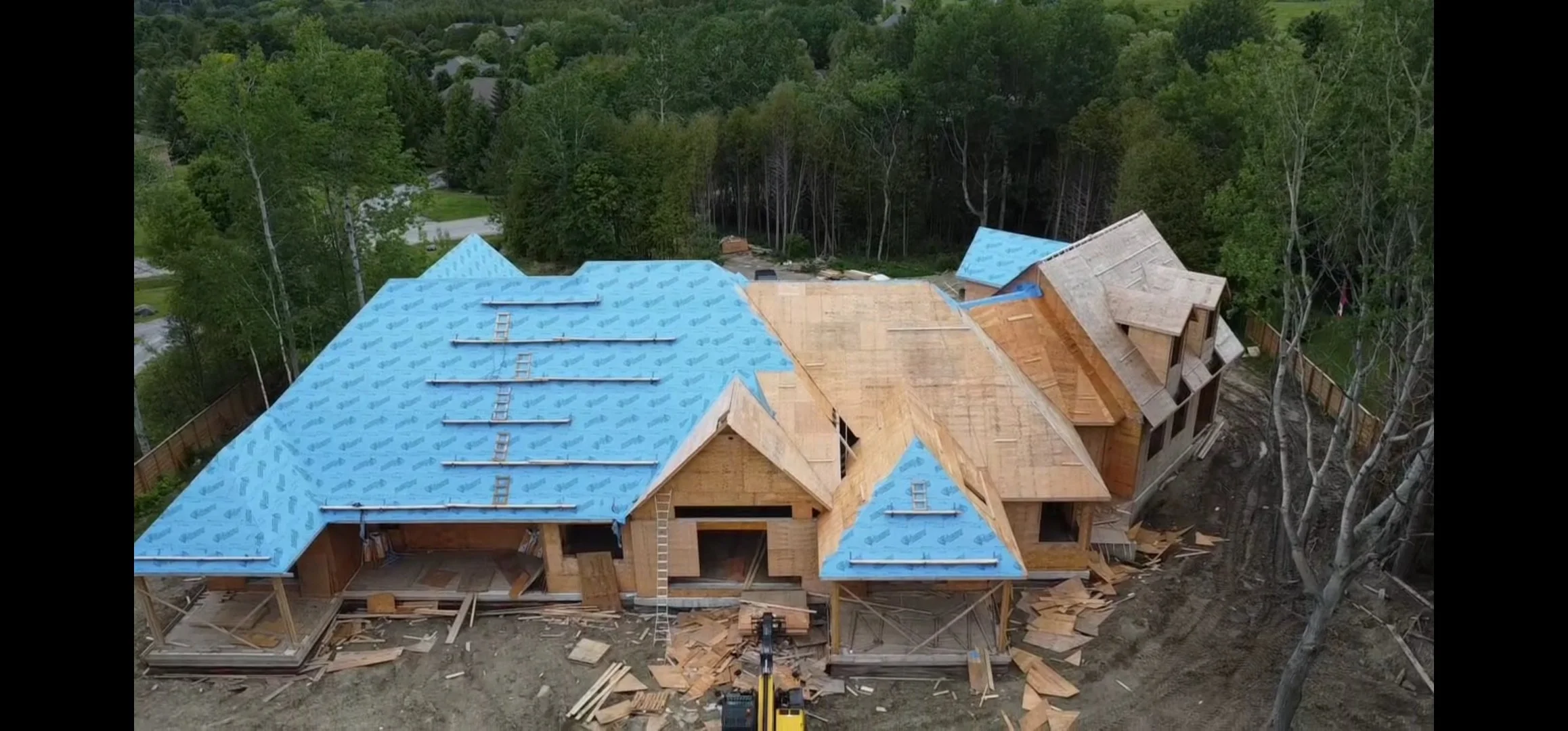 A house under construction with blue roofing underlayment, wooden walls, and partially completed roof sections surrounded by trees and construction debris.