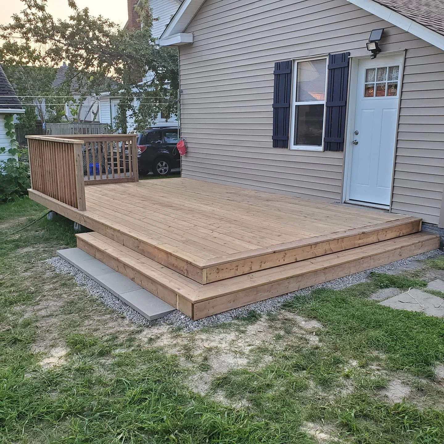 Newly built wooden deck with stairs outside a house, featuring a small railing on one side and a white door and window with blue shutters.