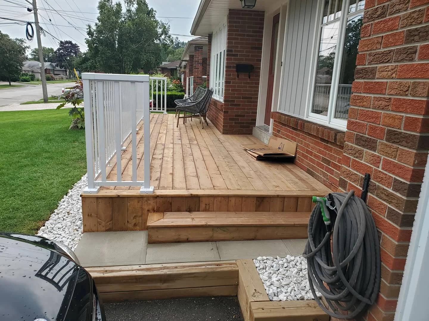 Newly built wooden porch with small steps, white railing, and a concrete patio in front of a brick house.