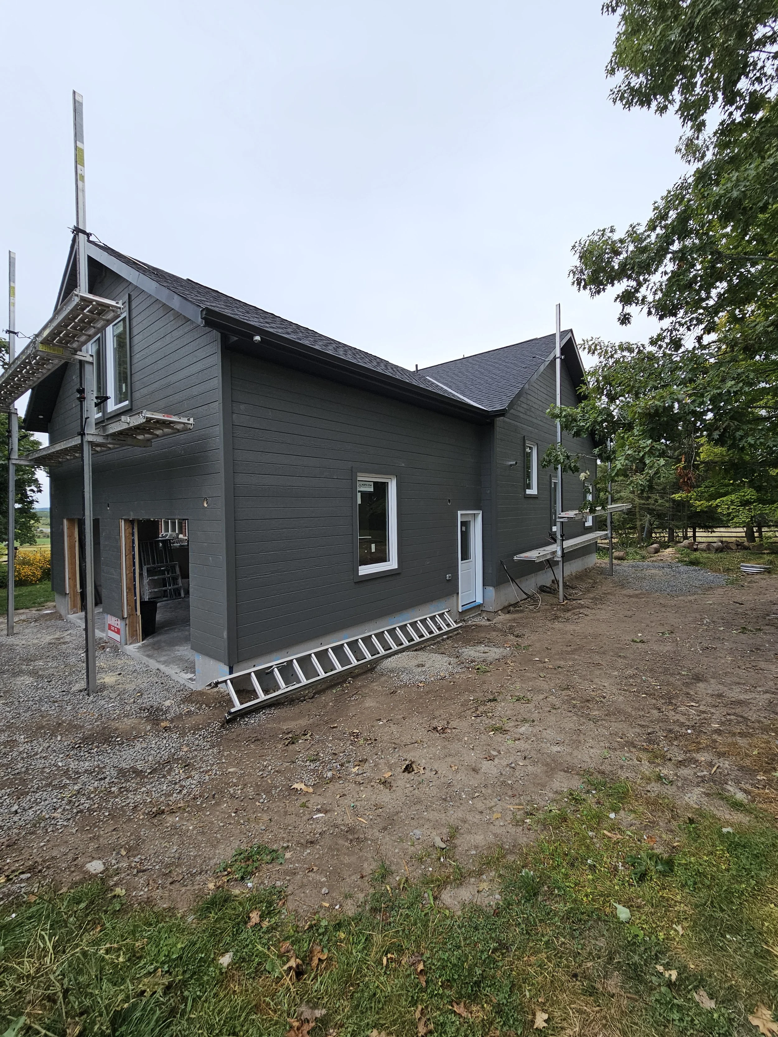 Side view of a house under construction with dark gray siding, multiple windows, a door, and scaffolding around it, on a partly cloudy day.