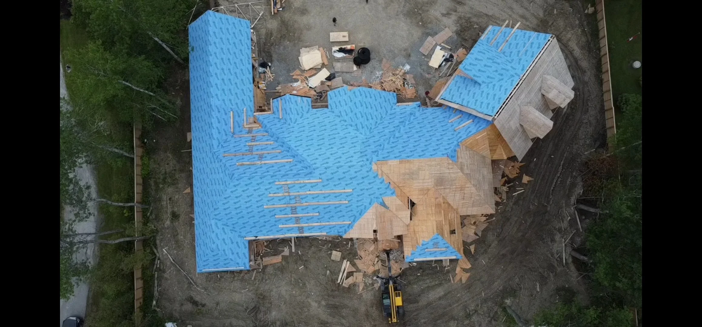  aerial view of a house under construction with a blue roofing underlayment, surrounded by construction materials and equipment, with trees and a dirt driveway nearby.