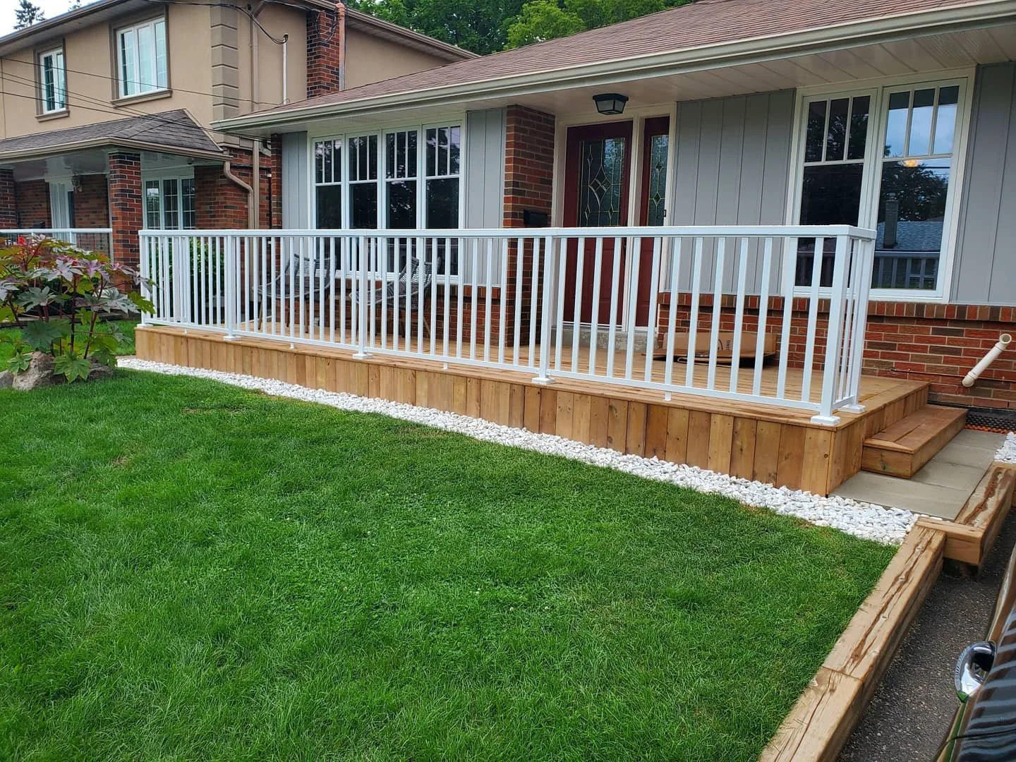 A backyard with a wooden deck, white railing, and green lawn. There are steps leading to the deck, and a small patio area with patio furniture. The house has brick and siding exterior, with large windows and a red front door.