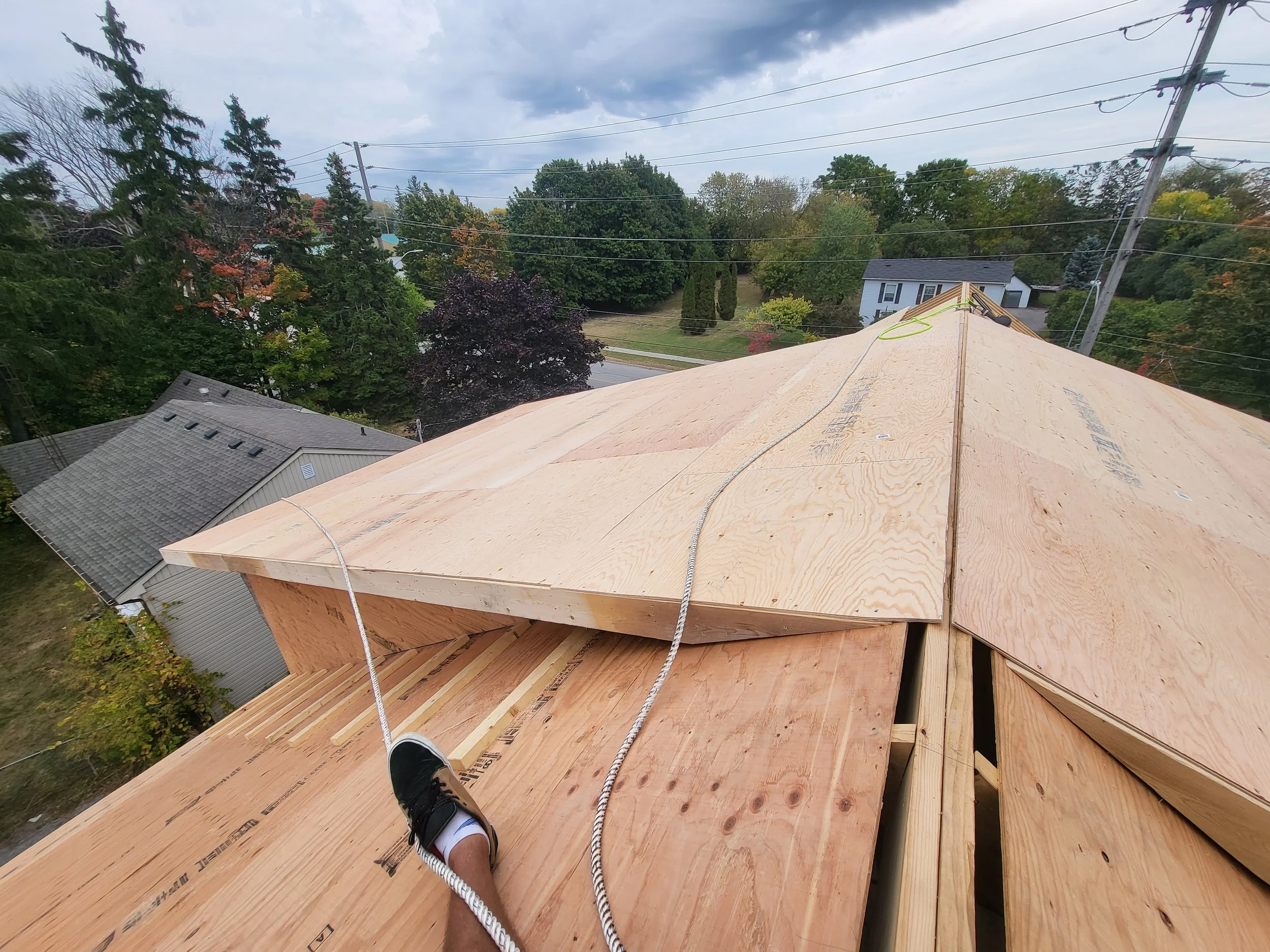 A view from a rooftop under construction, showing plywood sheathing, a person's foot in a black sneaker, and surrounding trees and houses.
