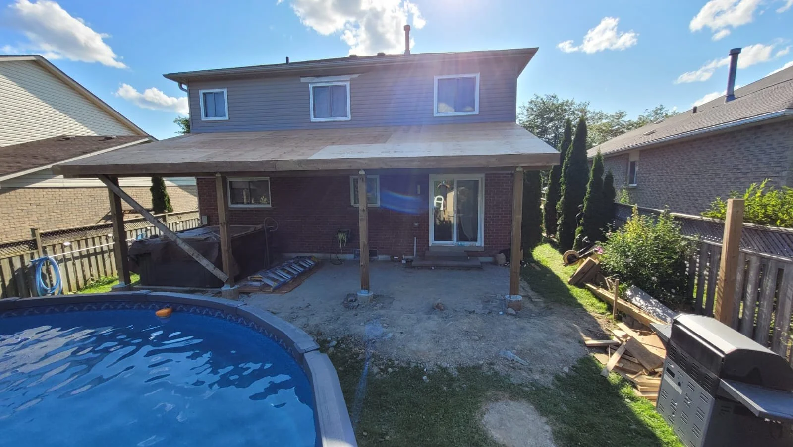 Backyard with a partially built covered patio, above ground pool in the foreground, safety equipment, and construction supplies. Neighboring houses on either side.