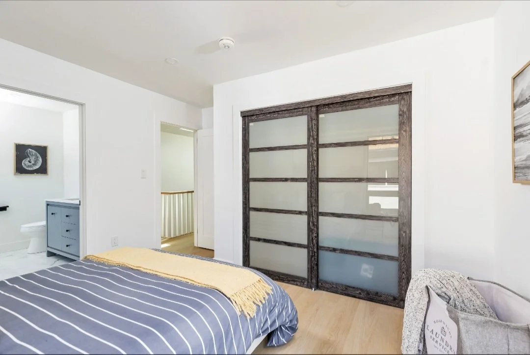 Bedroom with wooden sliding closet doors, a bed with a striped comforter and yellow throw, a gray laundry basket, and a doorway to a bathroom.