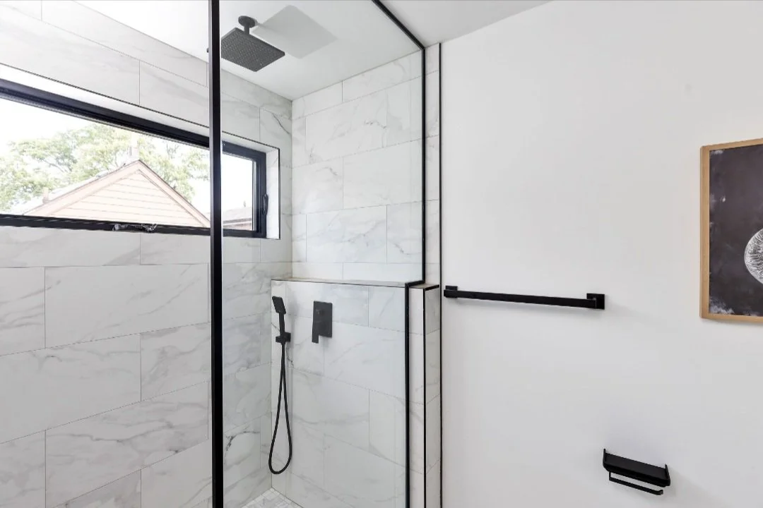 Modern walk-in shower with black fixtures, white marble tiles, and a window showing a house and trees outside.