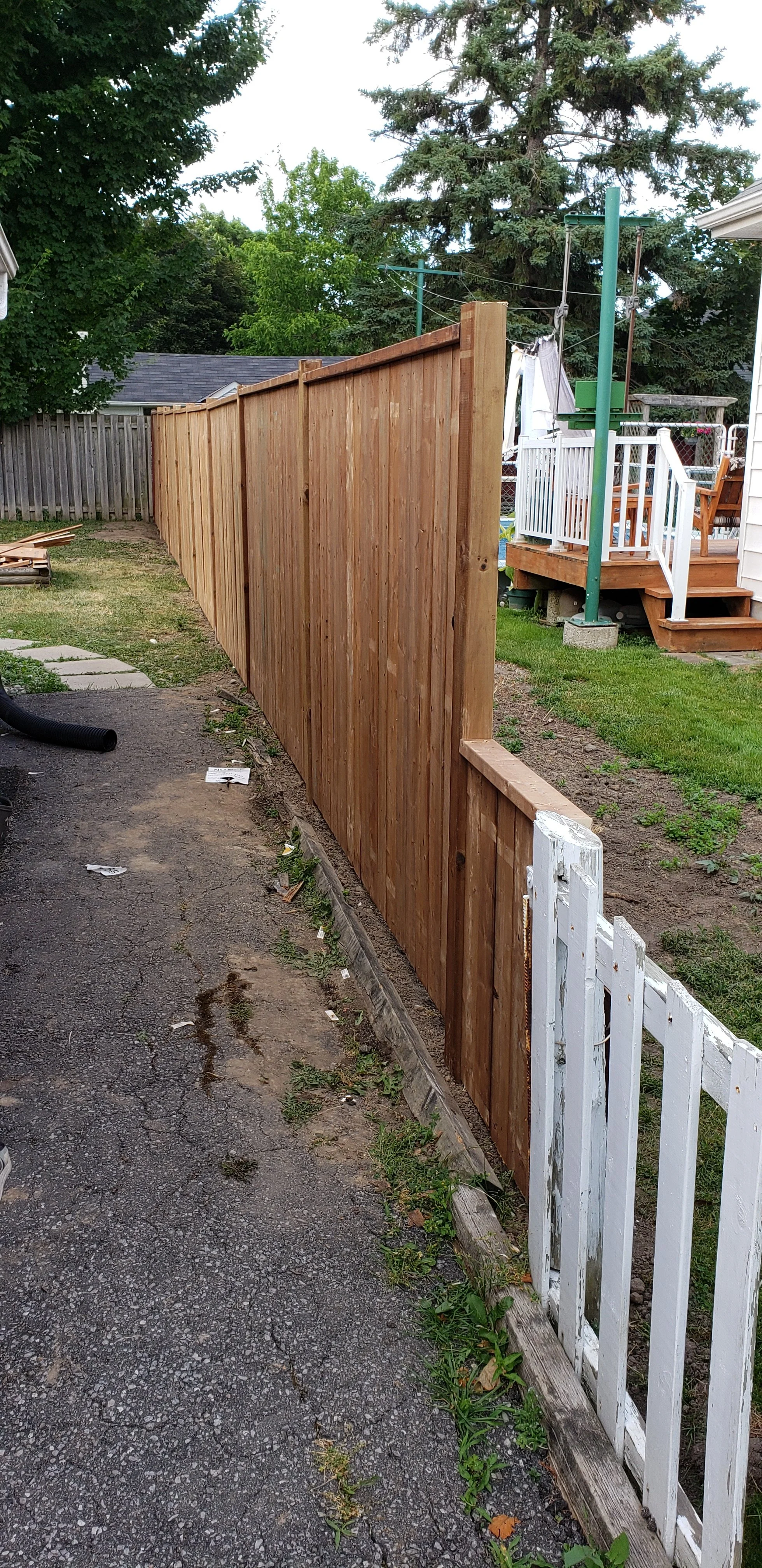 New wooden privacy fence being installed along a backyard, with an existing white picket fence and backyard deck visible.