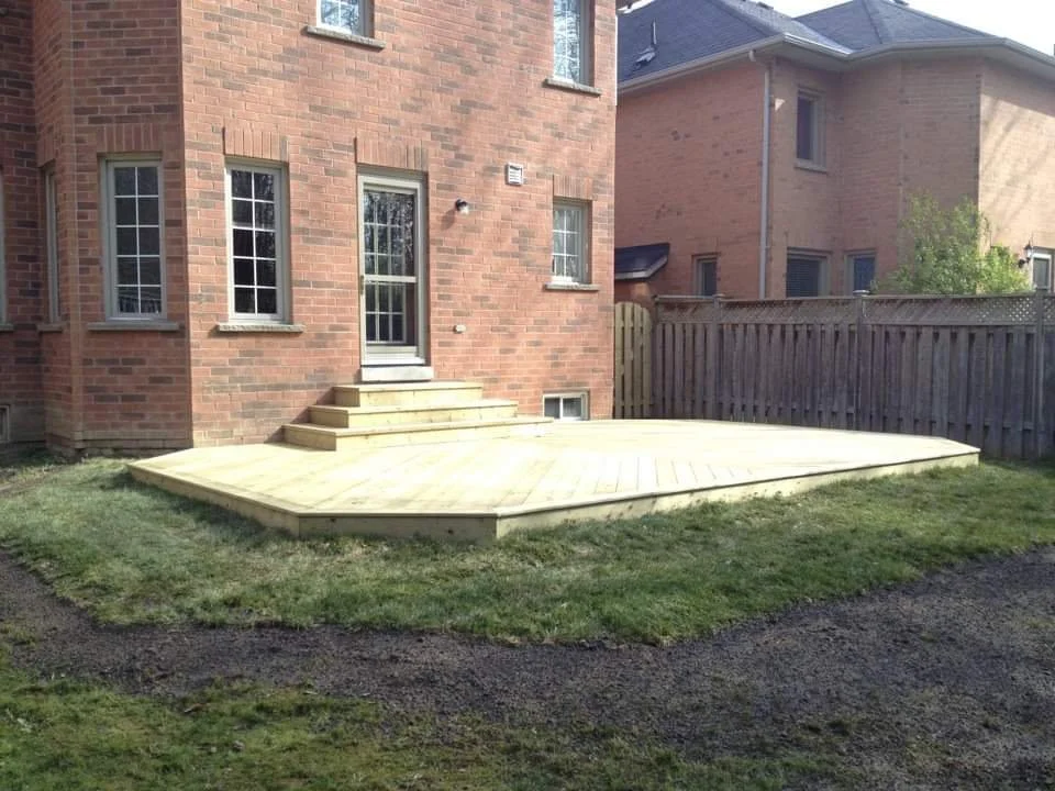 Newly built wooden deck with steps leading to a brick house, surrounded by a grassy yard and a wooden fence in the background.
