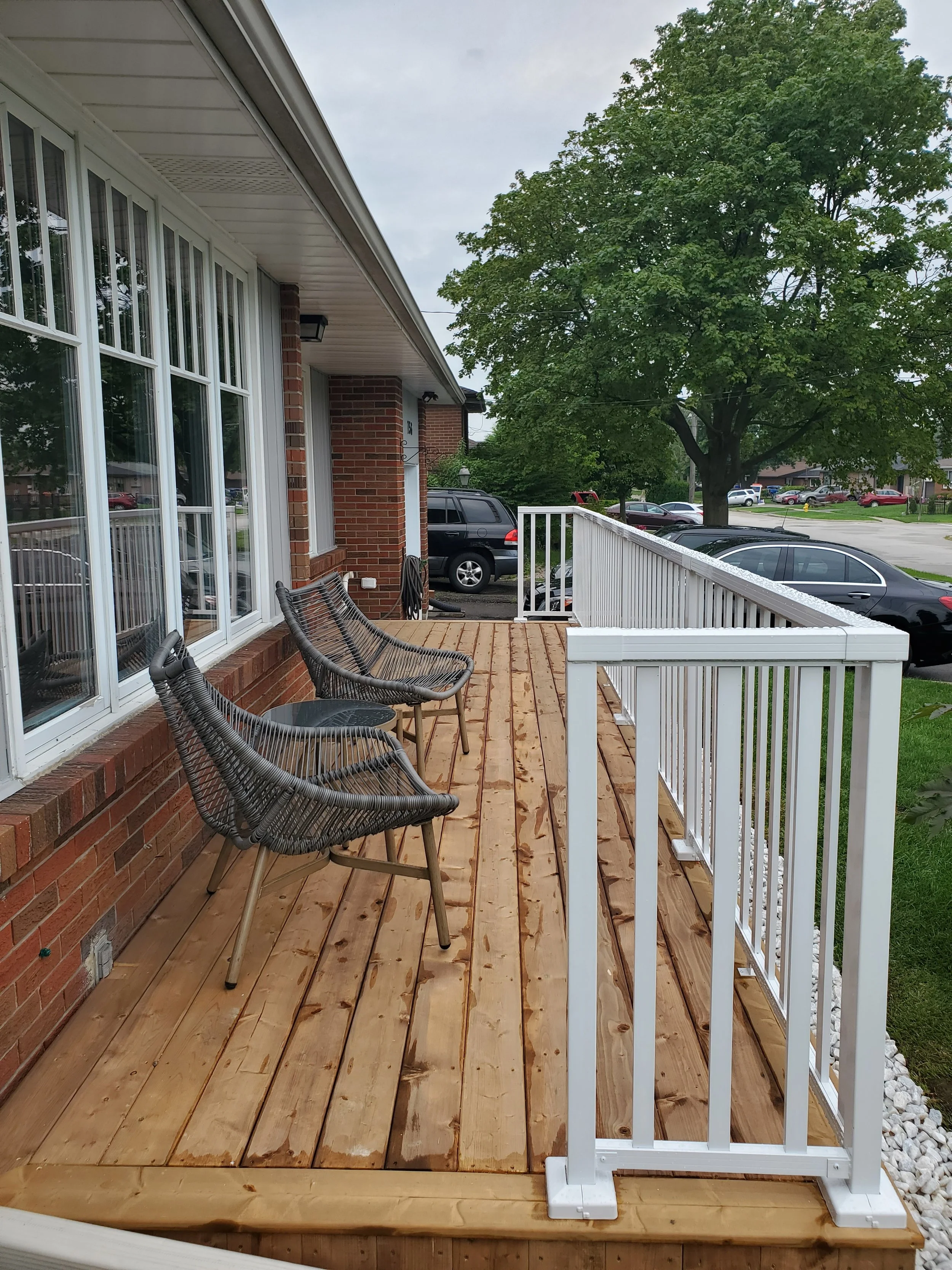 A newly built wooden deck with four chairs and an outdoor table, attached to a brick house with large windows, overlooking a parking lot with cars and a large green tree.