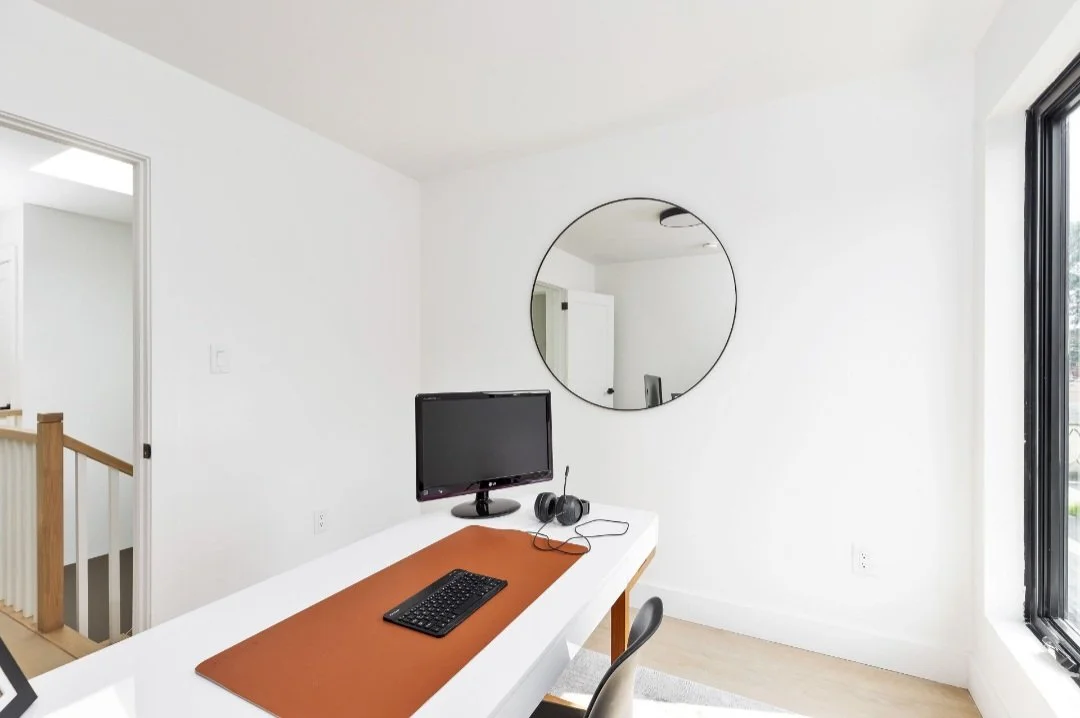 Home office desk with computer monitor, keyboard, headphones, mirror on white wall, window with sunlight