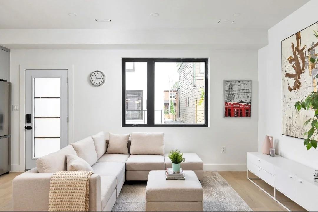 Bright living room with a white sectional sofa, a round wall clock, a large window, abstract wall art, a white media console, and a potted plant.