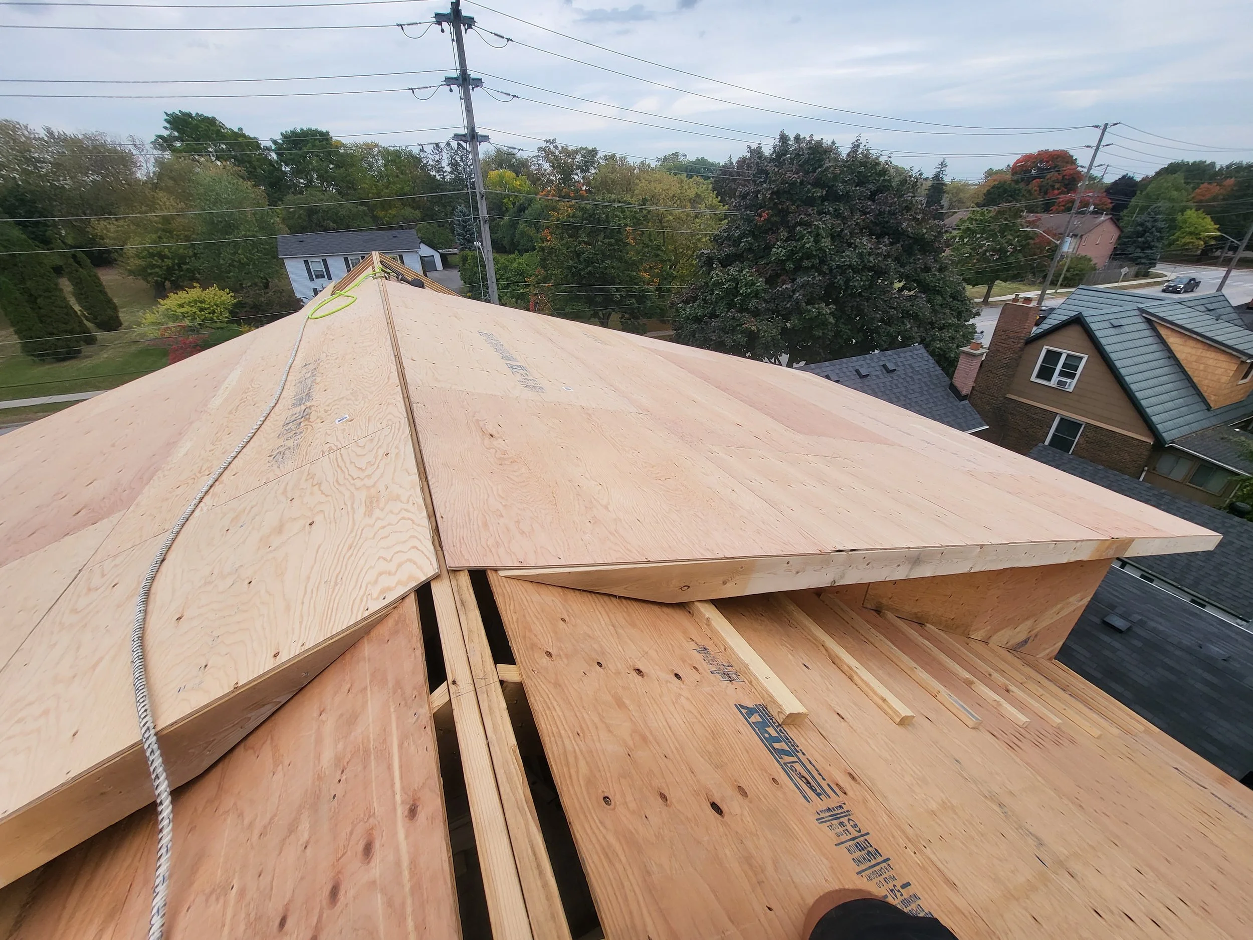 Construction site on a residential rooftop showing plywood sheathing and rafters with trees and houses in the background.