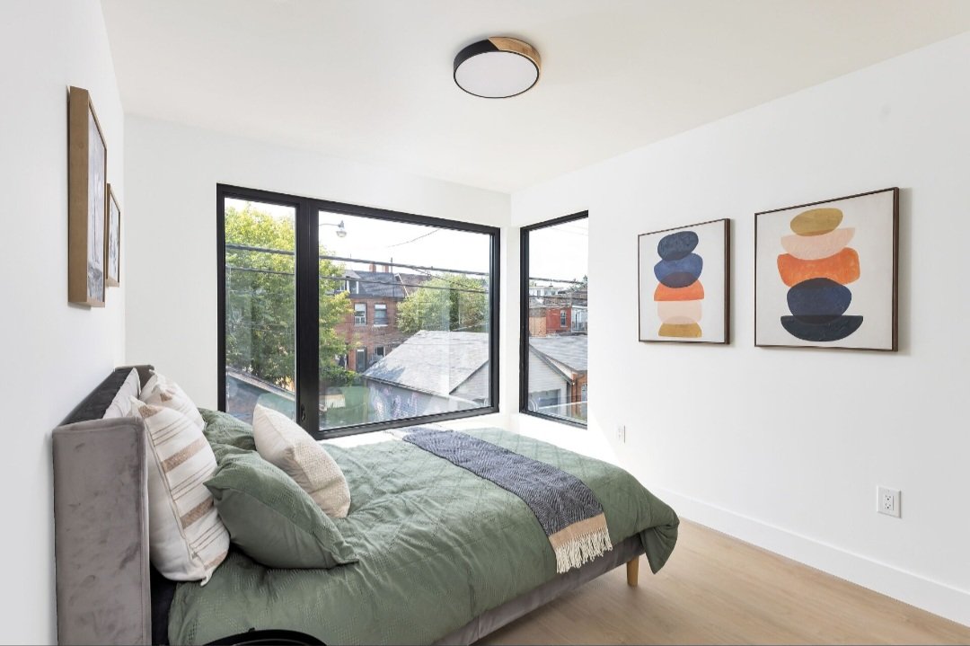 Bright bedroom with large corner window, green bedding and decorative pillows, modern artwork on white walls, wooden floor, ceiling light fixture.