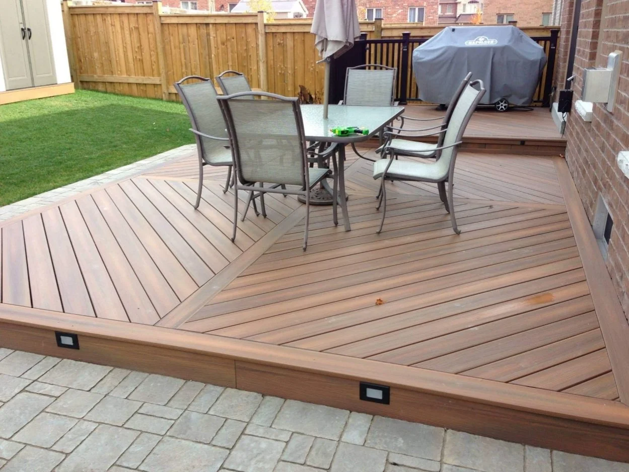 A backyard patio with a wooden deck, outdoor dining table with six chairs, an umbrella, a grill covered with a protective cover, and a small grassy area.