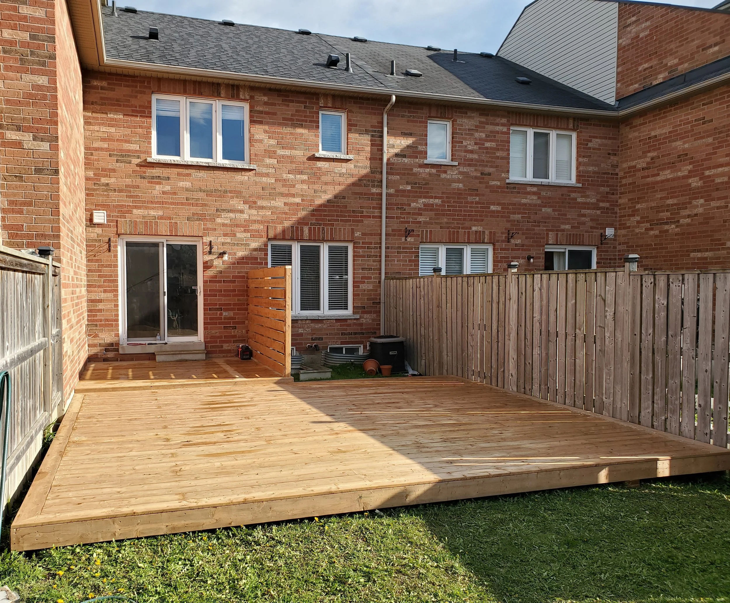 Newly built wooden deck in a backyard attached to a brick house with multiple windows, fenced on three sides with a small gate, and a sliding glass door leading inside.