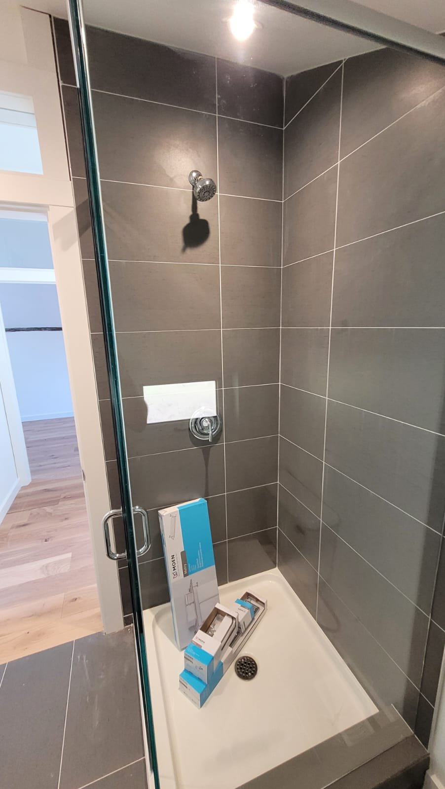A newly installed shower with gray tile walls, a chrome showerhead, and a glass door. There are boxes for a faucet and shower fixtures on the shower base, which has a drain and a small ledge at the bottom.