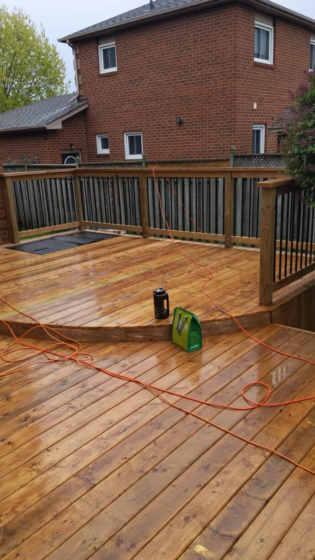 A wooden deck with wet planks and orange extension cords, a black lantern, a pack of weed killer, and a black doormat, surrounded by a wooden railing with black spindles in a backyard, with a brick house in the background.