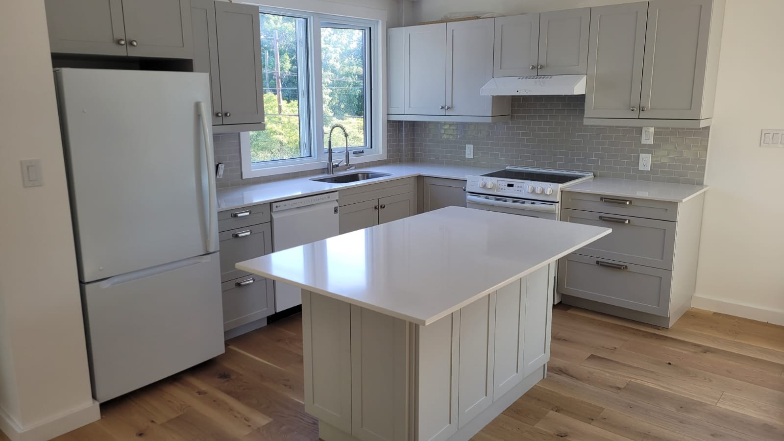 A modern kitchen with white cabinets and a white island, stainless steel appliances, a window above the sink, and hardwood flooring.