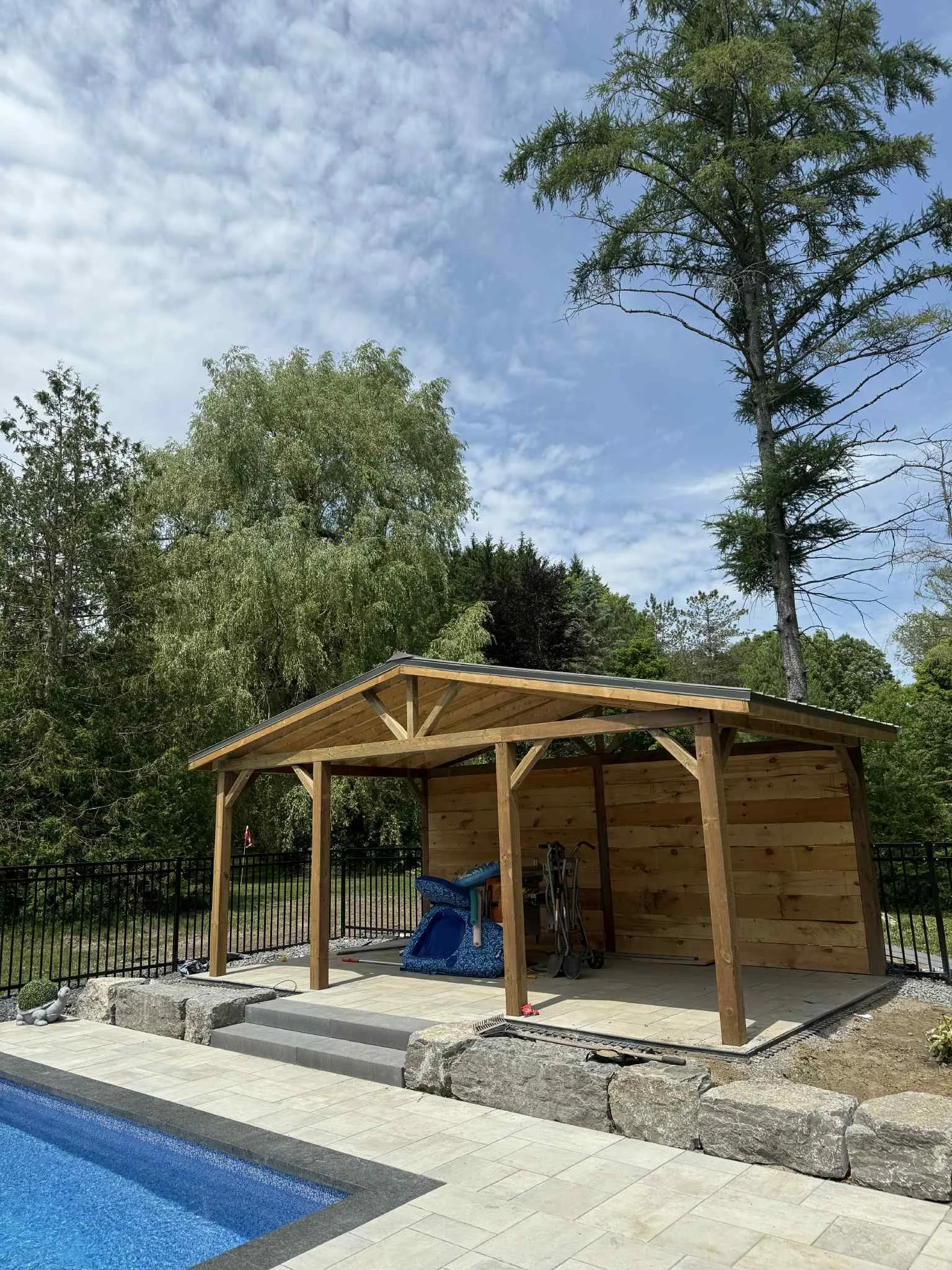 A backyard scene featuring a partially constructed wooden poolside shelter with a sloped roof, next to a swimming pool, surrounded by trees and a fence.