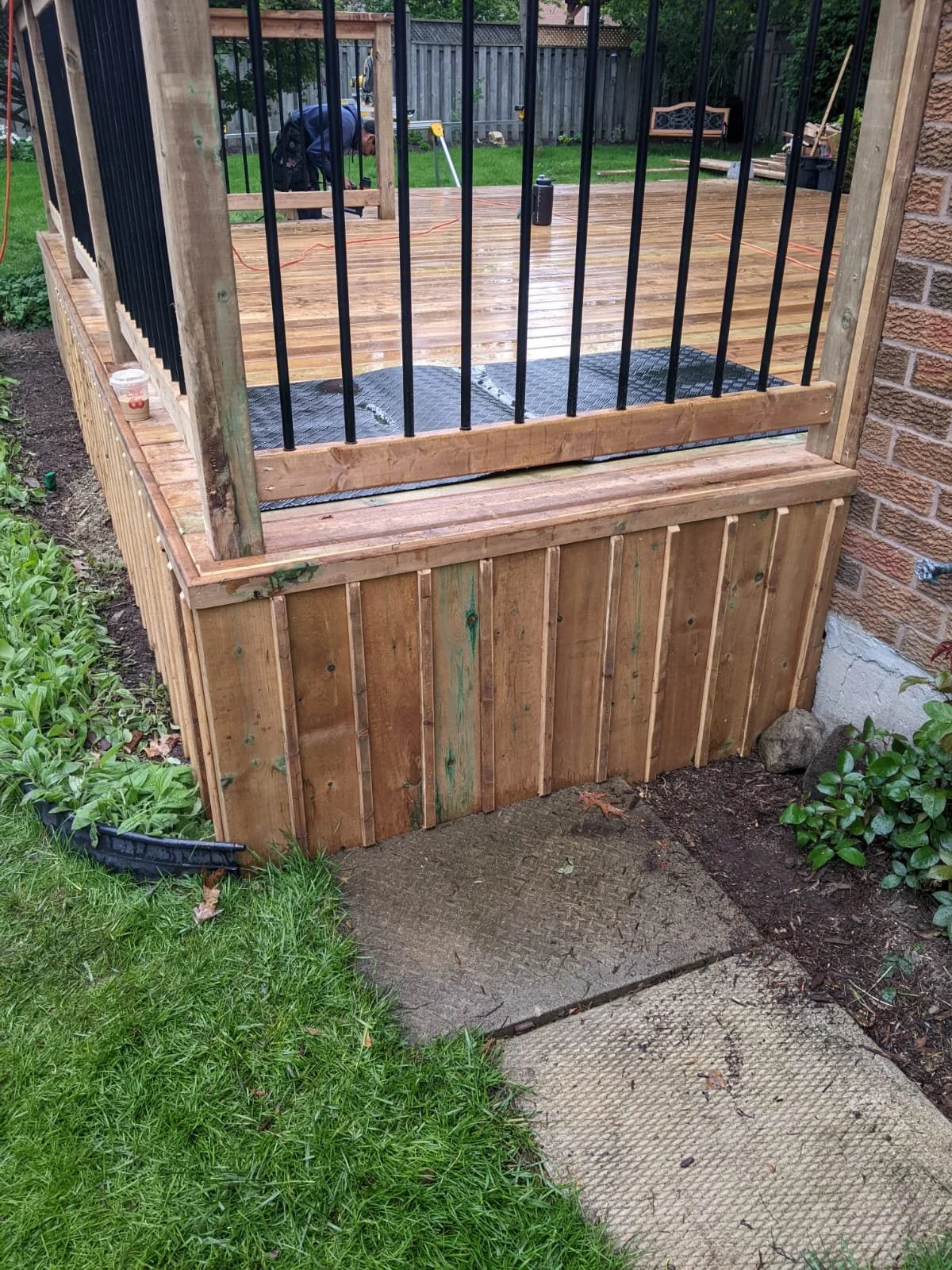 Newly built wooden deck with black metal railings next to brick wall, with a person working on the deck in the background, and garden plants and concrete path in the foreground.