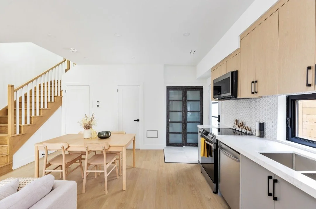 Modern kitchen and dining area with light wood cabinets, stainless steel appliances, a farmhouse door, and a staircase with a wooden banister.