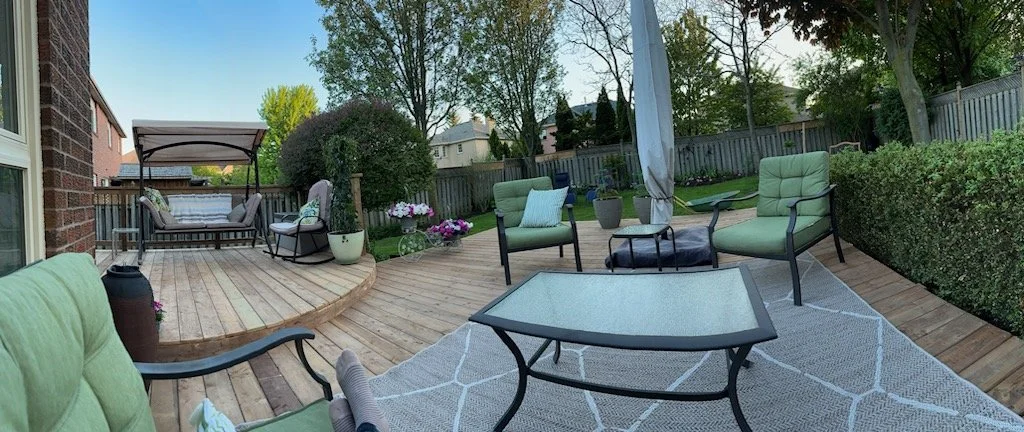 Backyard patio with green cushioned outdoor chairs, a glass coffee table, potted plants, a wooden deck, a covered swing, and a white umbrella, surrounded by a high fence and trees.