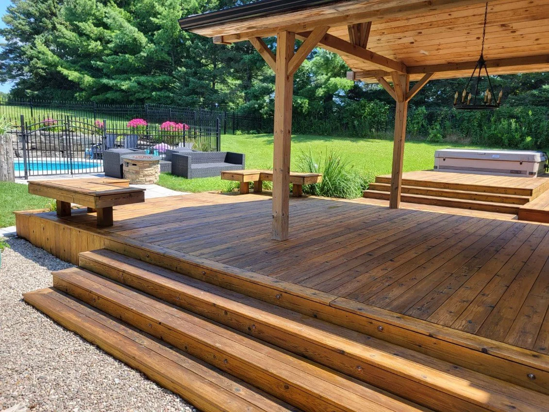 Wooden backyard deck with stairs, covered porch, outdoor furniture, hot tub, and swimming pool in a grassy yard with trees in the background.
