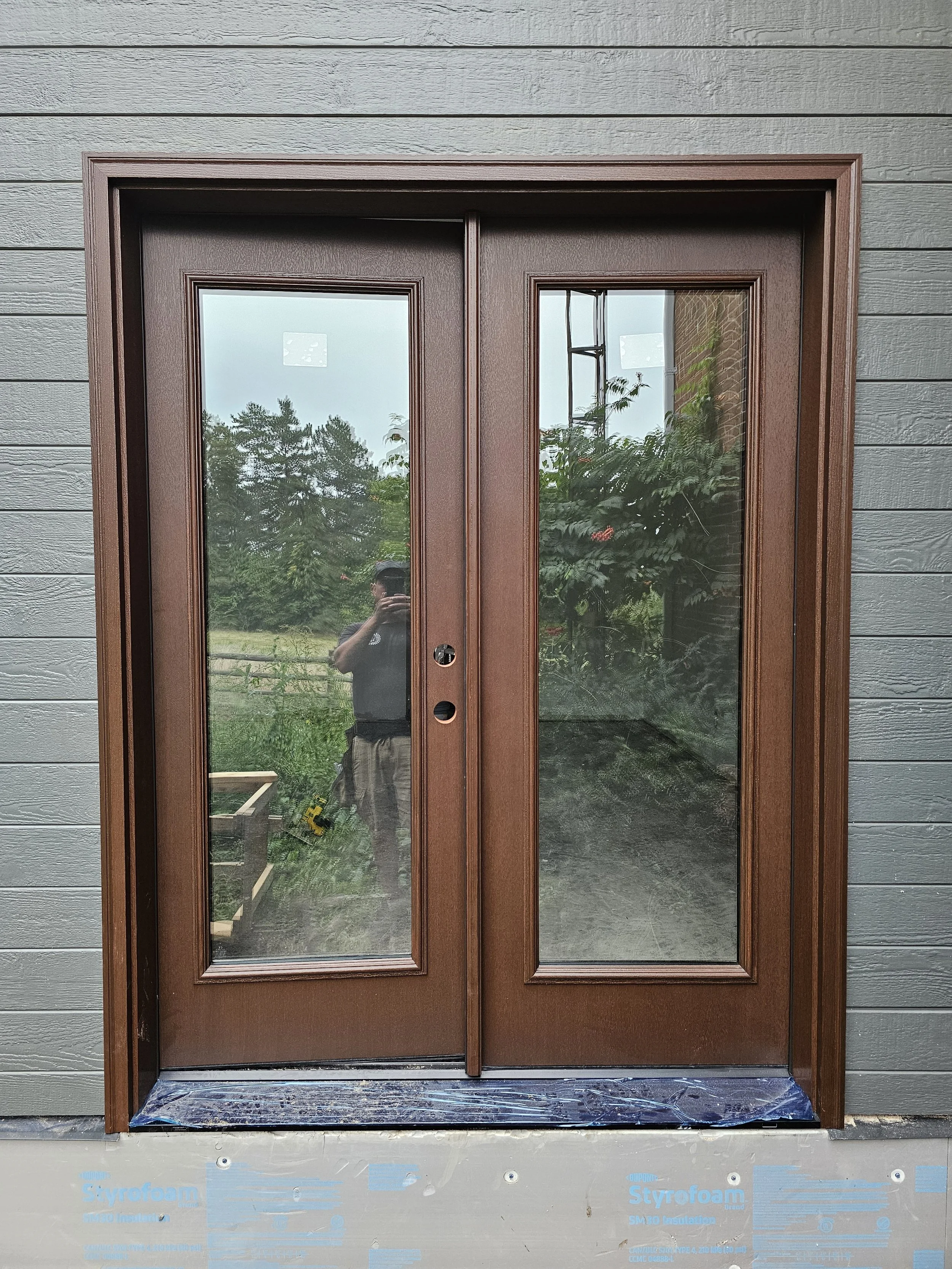 Double glass sliding door with brown frame installed on a house. The exterior wall has horizontal gray siding.