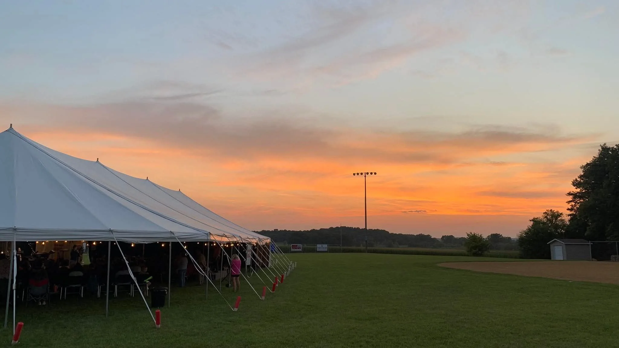 An outdoor scene at sunset with a large white event tent on the left, people gathered inside under the tent, and trees in the distance under a colorful sky.