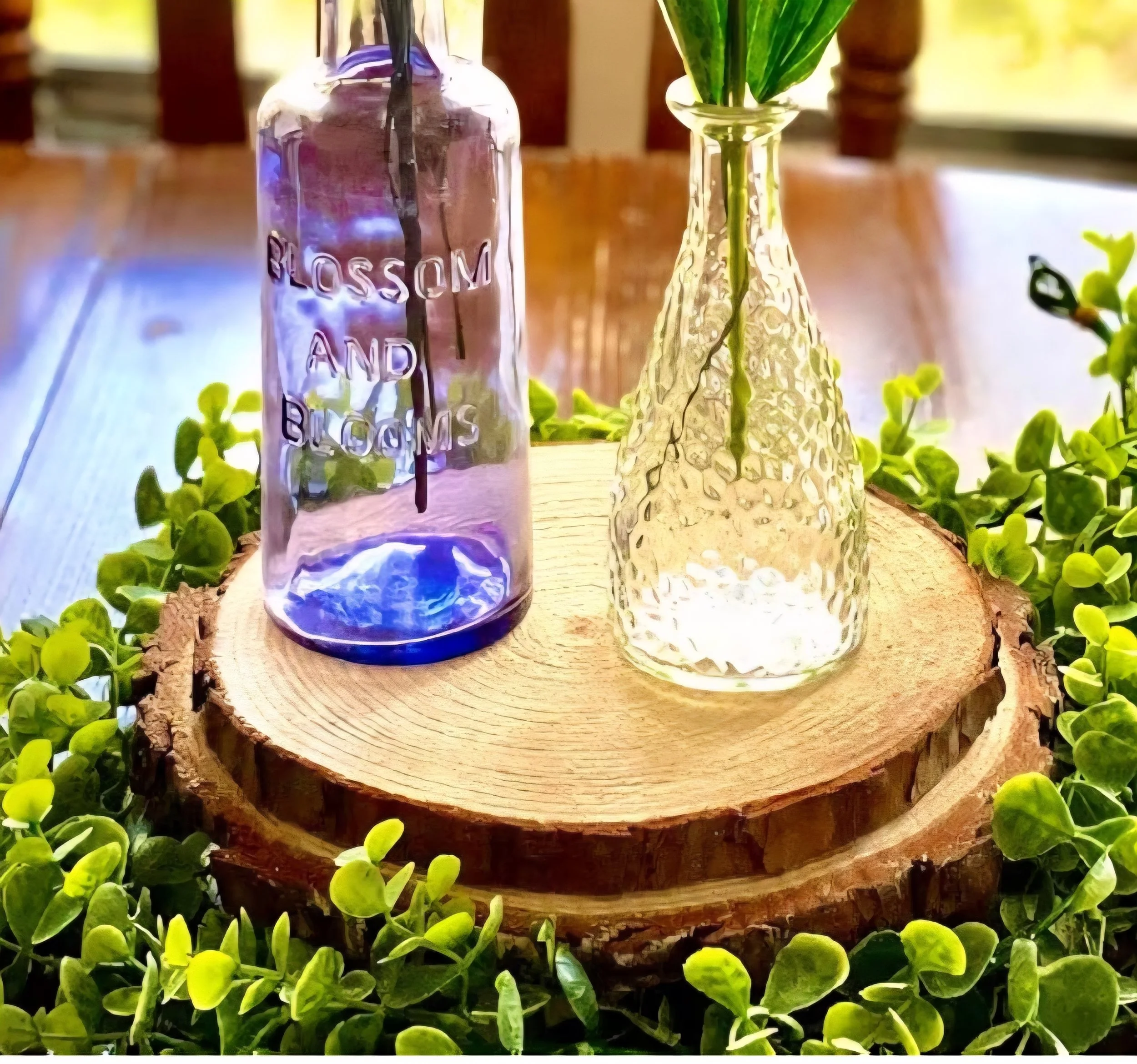 Two glass bottles placed on a round wooden slice, surrounded by green foliage.