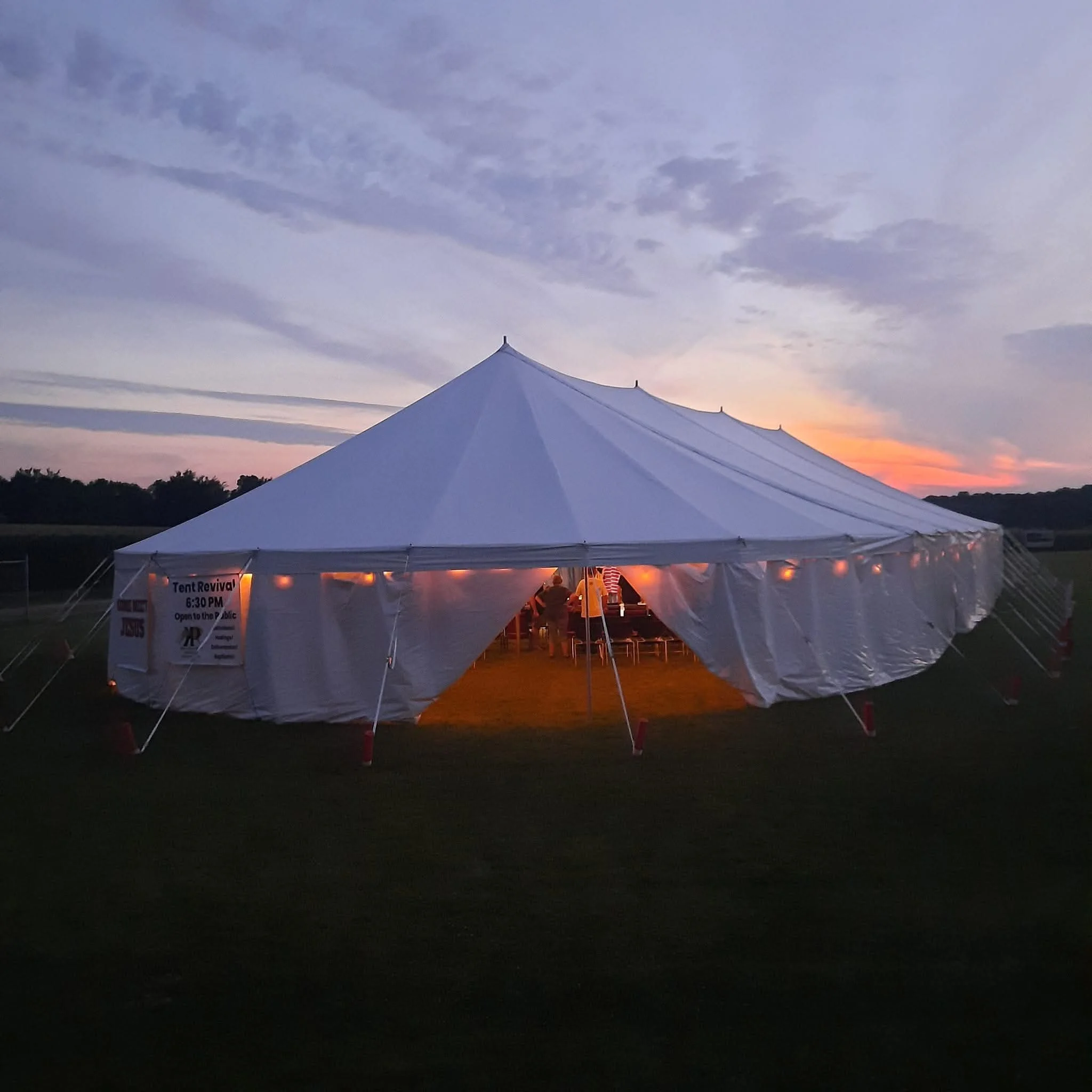 A large white event tent set up outdoors during sunset with string lights inside, with a sign on the side that reads "Tent Revival 6:30 PM open to the public."