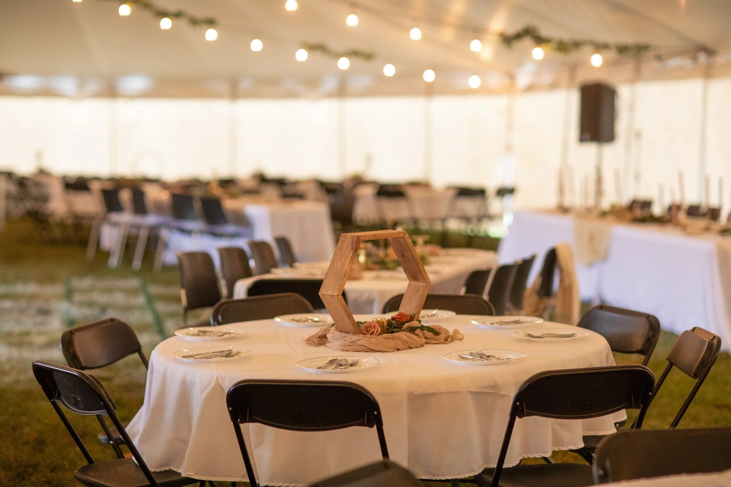 Decorated reception tent with round tables, white tablecloths, and black chairs, featuring centerpieces with floral accents, string lights, and a long buffet table in the background.