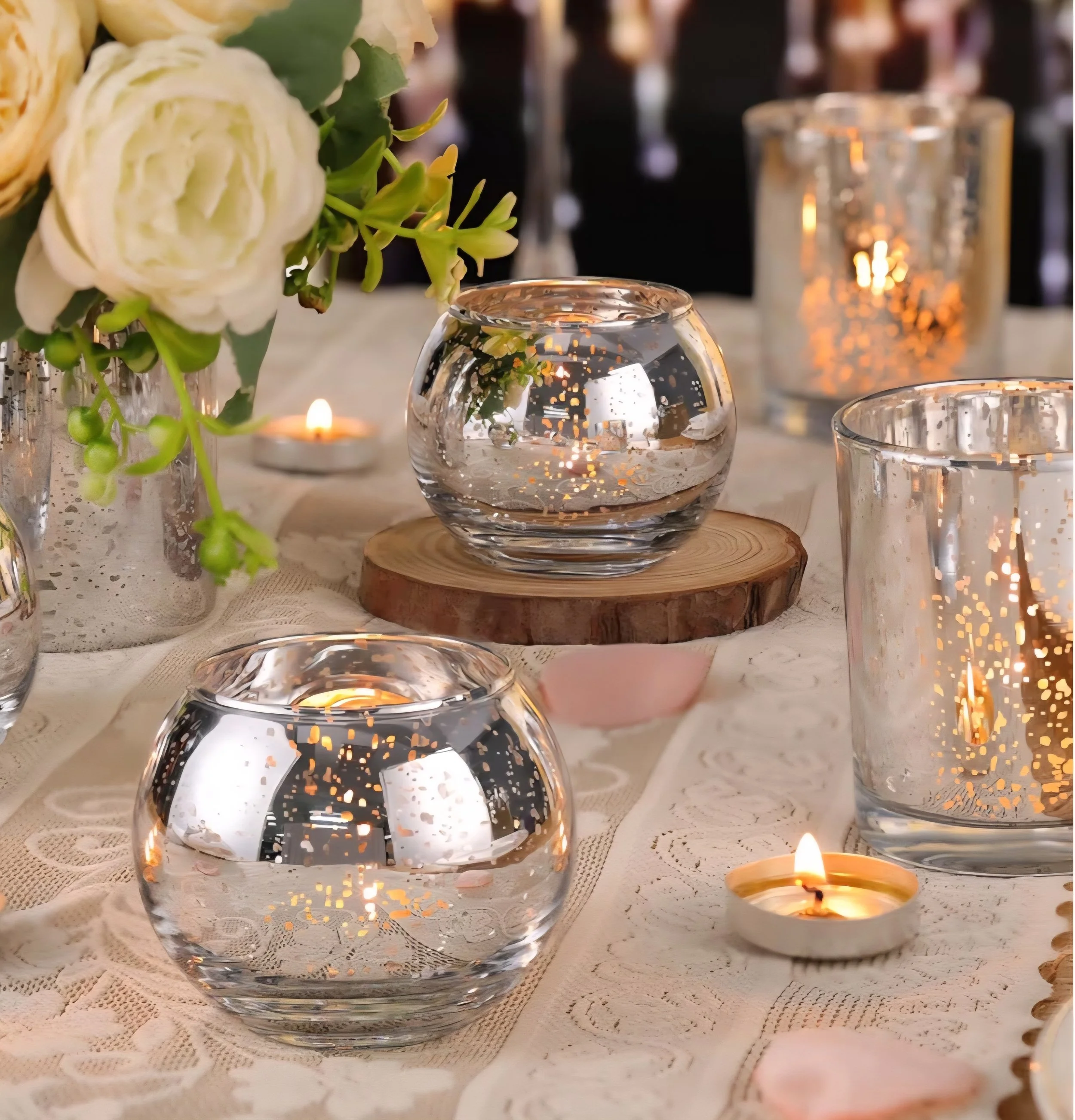 Decorative table setting with glass candle holders, lit candles, floral arrangements, and lace tablecloth.