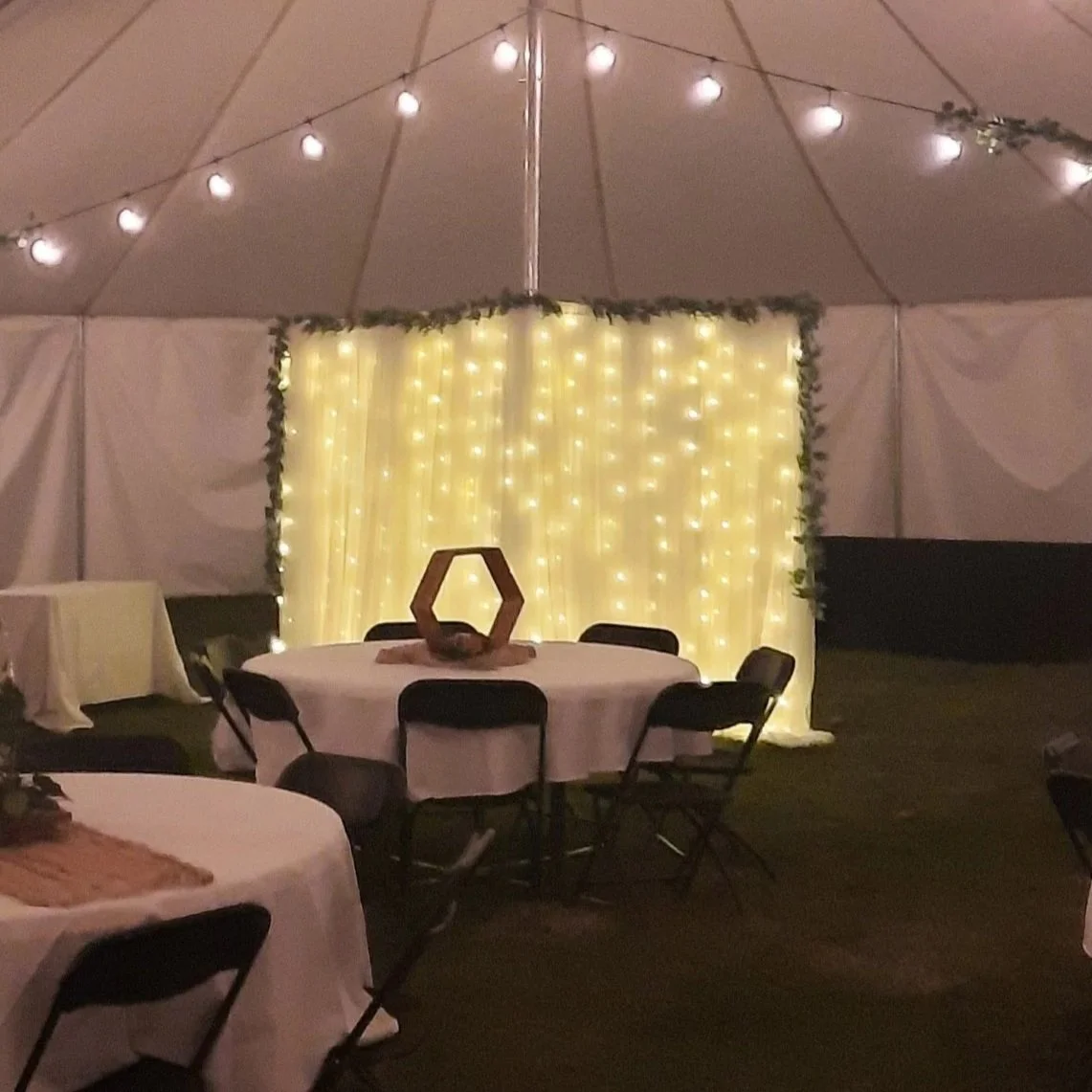 Event space with round tables covered in white tablecloths, black chairs, a backdrop of warm fairy lights, and a hexagonal wooden centerpiece on the main table. The setup is inside a large tent with string lights along the ceiling.