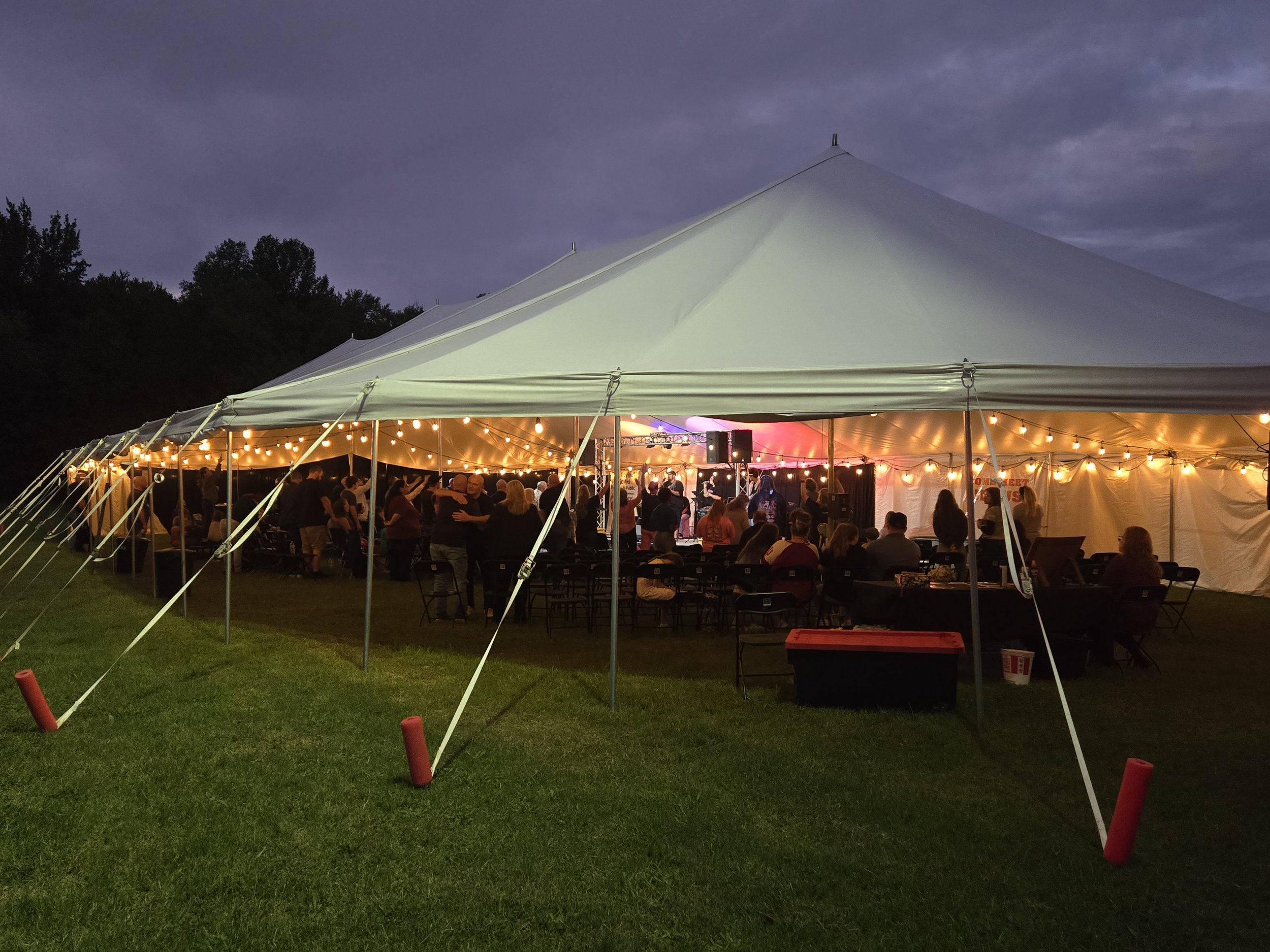 Outdoor event with a large white tent decorated with string lights, where people are gathered inside at dusk.