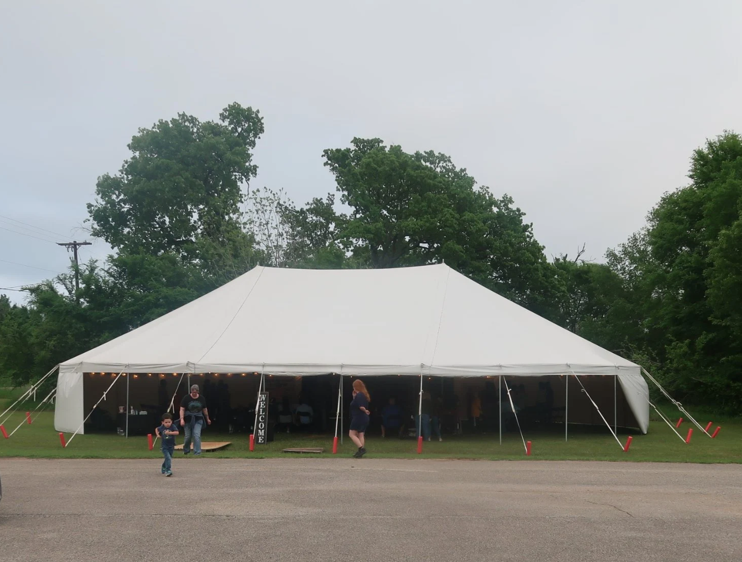 A large white event tent set up outdoors with people inside and outside, on a grassy area with trees in the background and a cloudy sky.