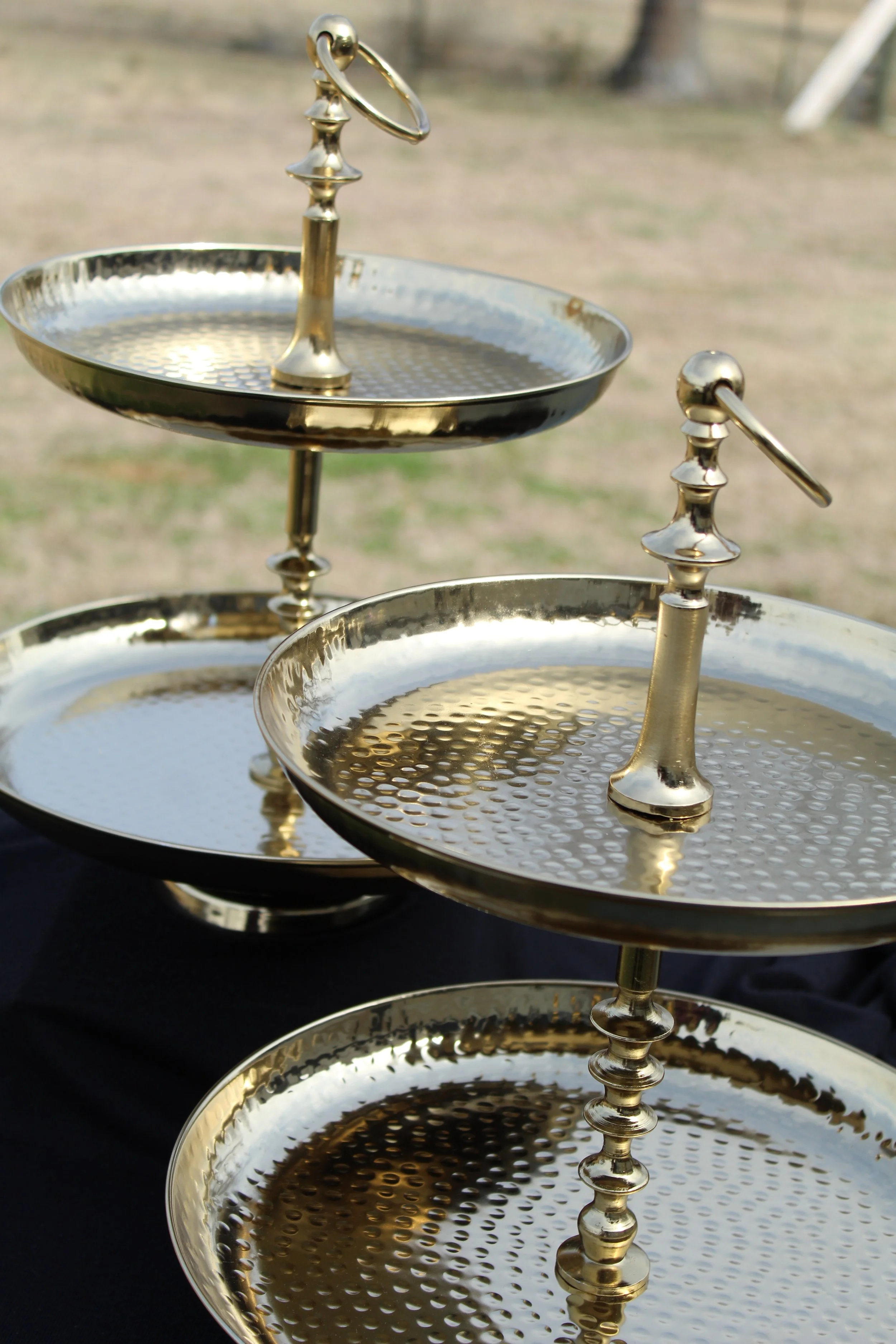 Three-tier metallic serving trays with ornate handles, set outdoors on a black surface, against a blurred natural background.