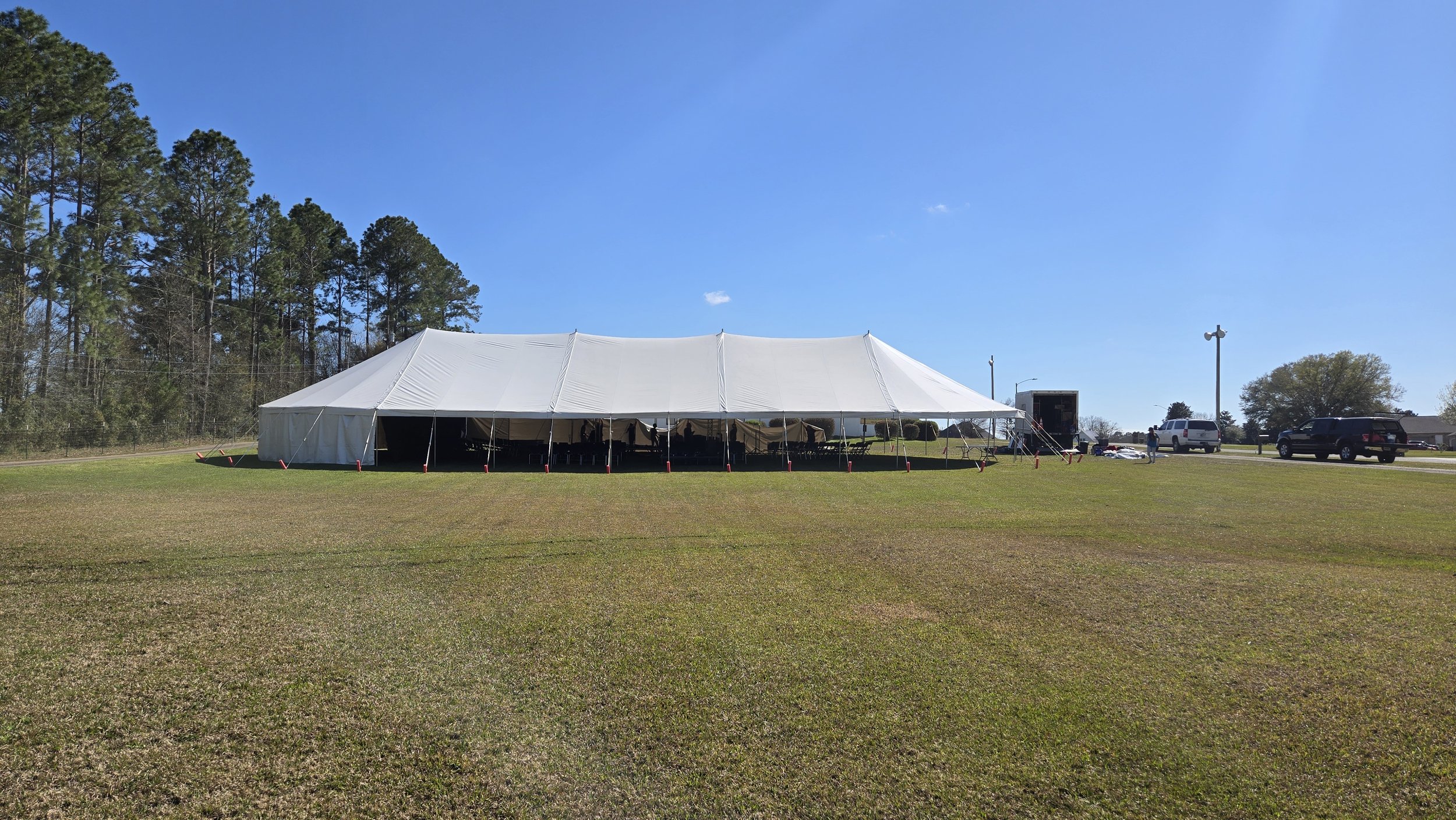 Large white event tent set up on a grassy field with cars and people in the background, clear blue sky.