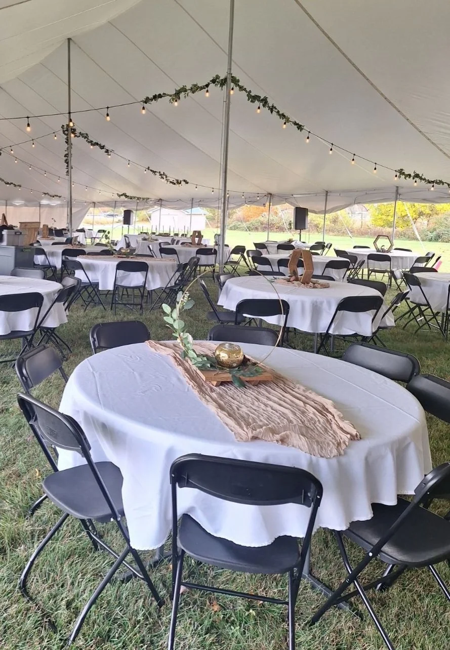 Wedding reception setup inside a large white tent with decorated tables, string lights, greenery, and natural outdoor scenery in the background.