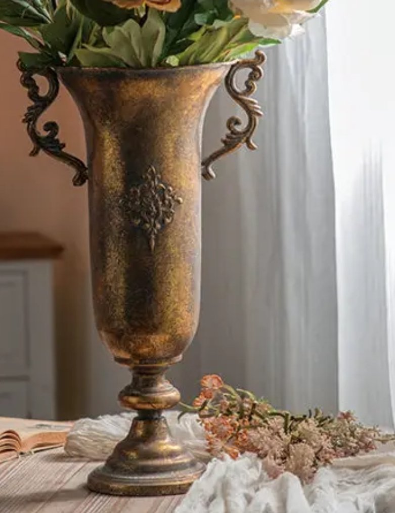 A tall ornate gold and bronze vase with decorative handles, holding white and pale pink flowers, placed on a table with a cloth and some pink flowers at the base.