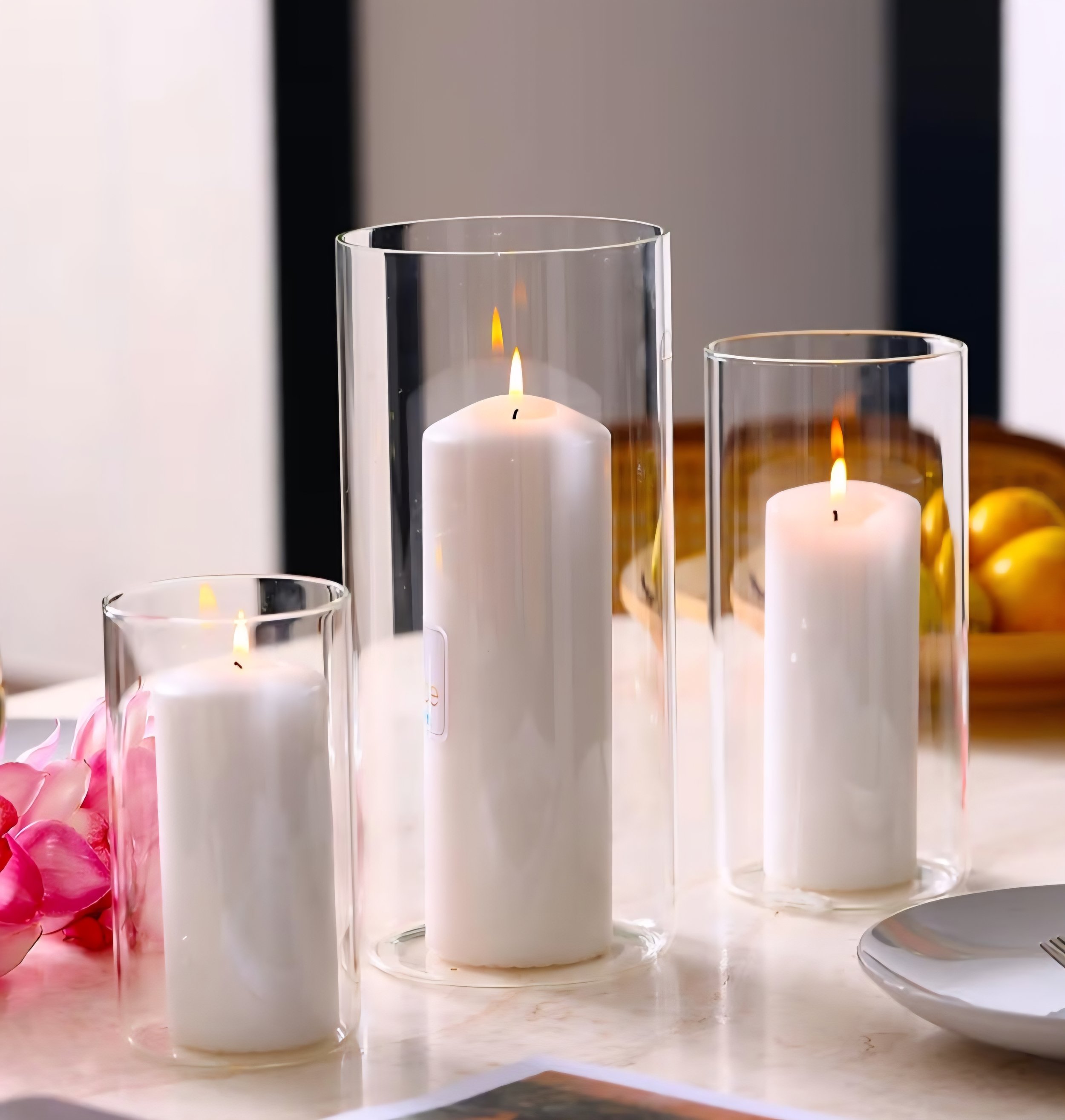 Three white candles burning inside tall, clear glass candle holders of varying heights on a decorated table.
