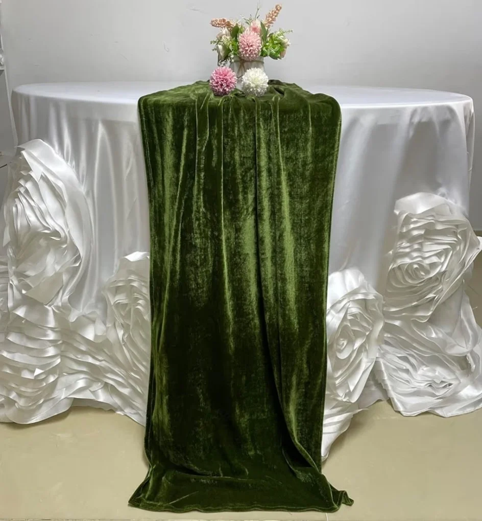 A round table with a white satin tablecloth and decorative white satin fabric roses, with a dark green velvet ribbon running down the center, topped with a floral arrangement of pink, white, and purple flowers.