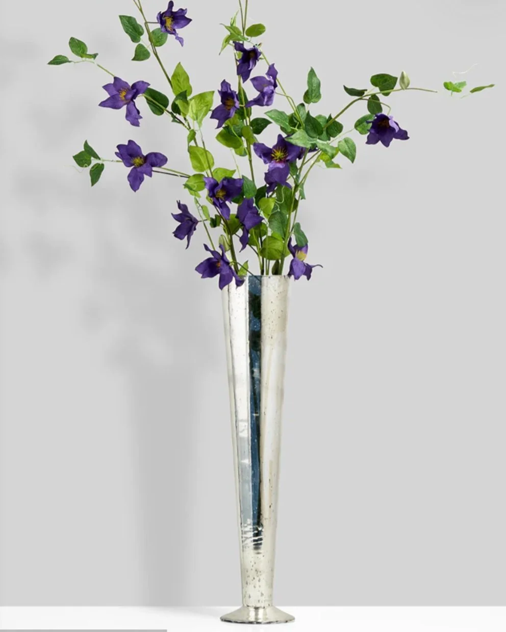 A tall, silver, modern vase with a slim, elongated shape, filled with purple flowers and green leaves.