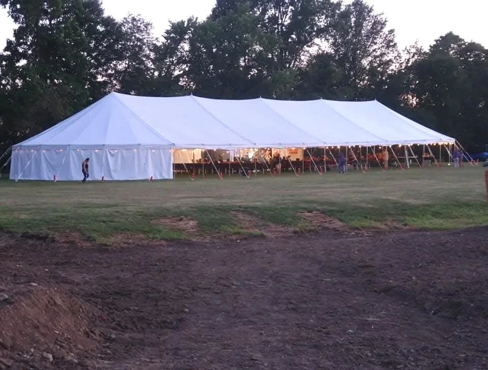 A large white event tent set up on a grassy field, with several people near it, and trees in the background during dusk or early evening.