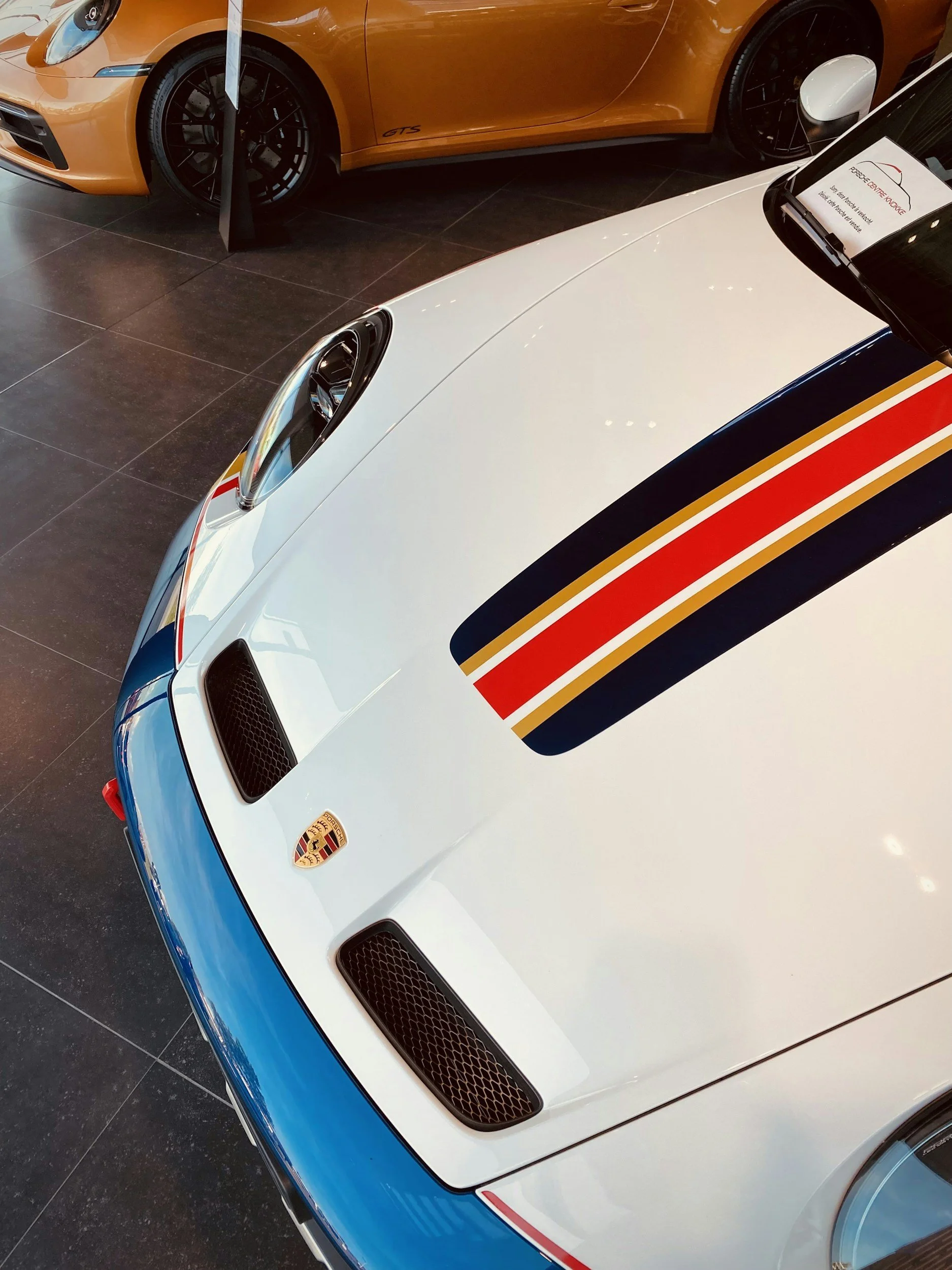 Close-up of a white Porsche sports car with a racing stripe in red, gold, and black on the hood, and the Porsche emblem visible. Part of an orange car is visible in the background.