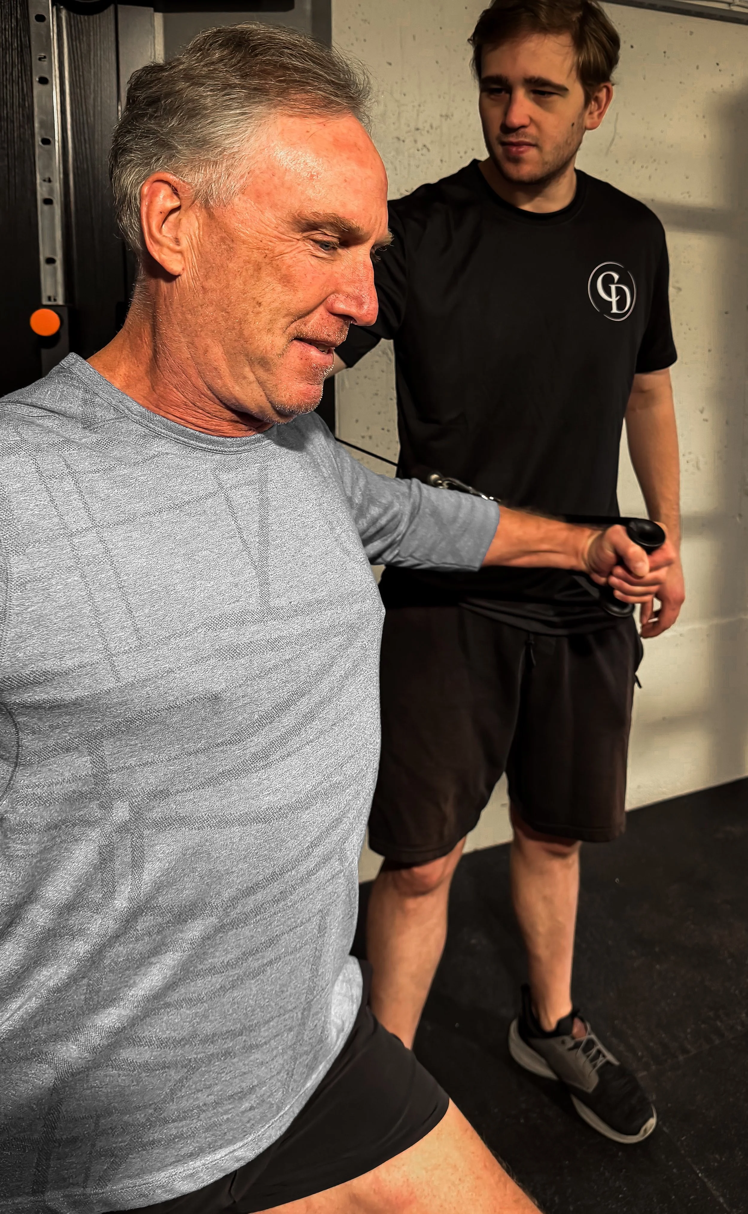 A senior man in a gray athletic shirt and black shorts is stretching at the gym, assisted by a younger man in a black t-shirt with a logo and black shorts, holding a dumbbell.