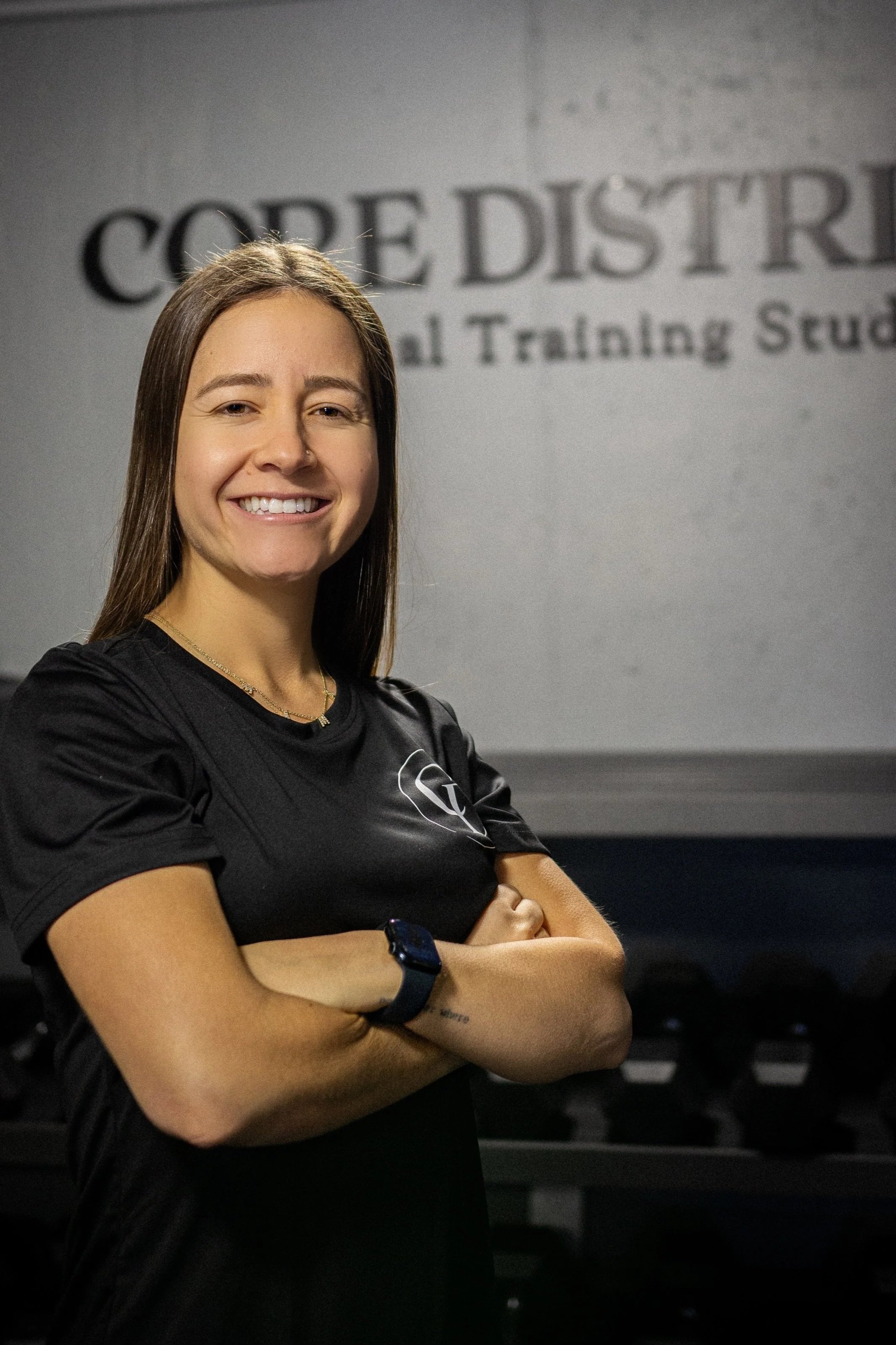 A smiling woman with brown hair wearing a black athletic shirt and a smartwatch, standing arms crossed in front of a wall with a logo and text related to a coaching or training school.