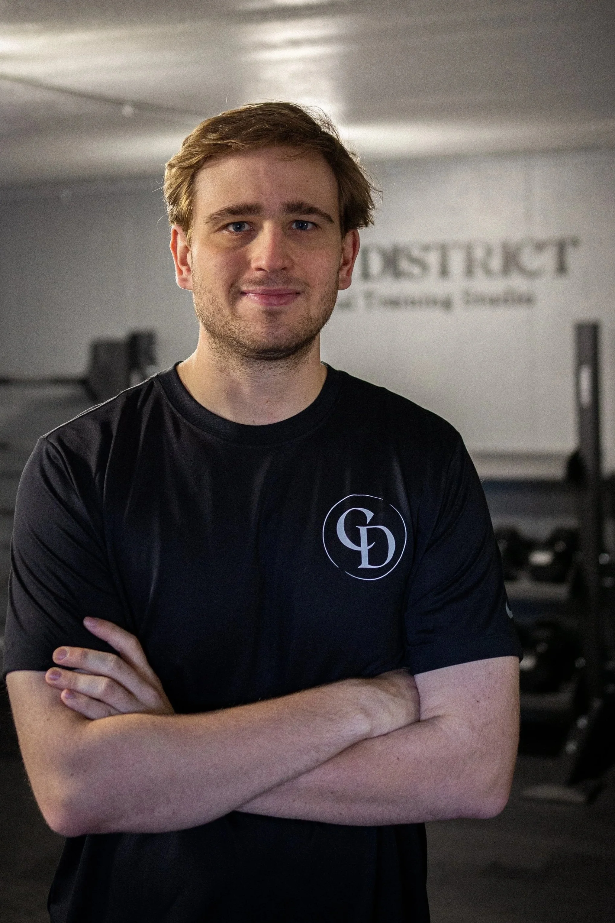 A young man with light brown hair and blue eyes, smiling, standing with arms crossed in a gym. He is wearing a black athletic shirt with a logo on the chest, and the gym background includes weightlifting equipment and a partial view of a sign that reads 'DSTRUCT'.