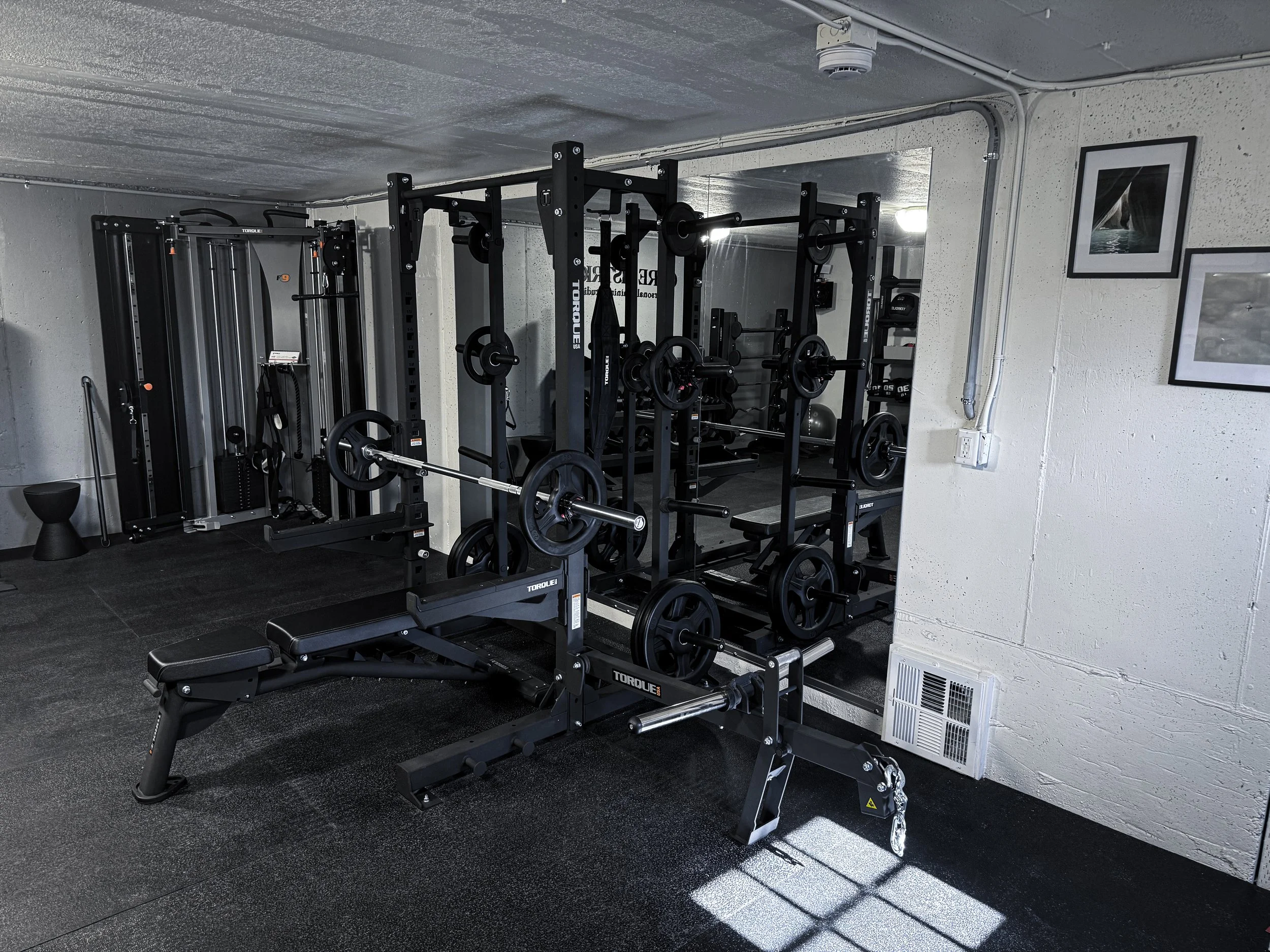A small gym room with weightlifting equipment including barbells, weight plates, and a bench in the foreground. There are framed photographs on the white brick wall and a ventilation grate near the floor.