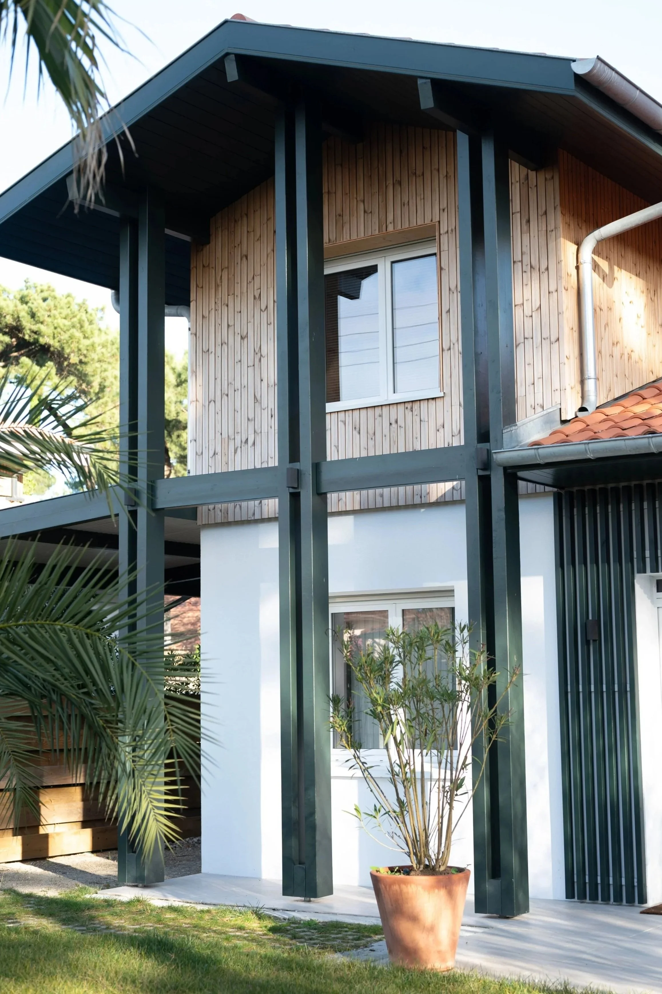 Maison moderne avec façade en bardage bois et poteaux en bois vert foncé, fenêtre au premier et au second étage, pot de plante devant la maison.